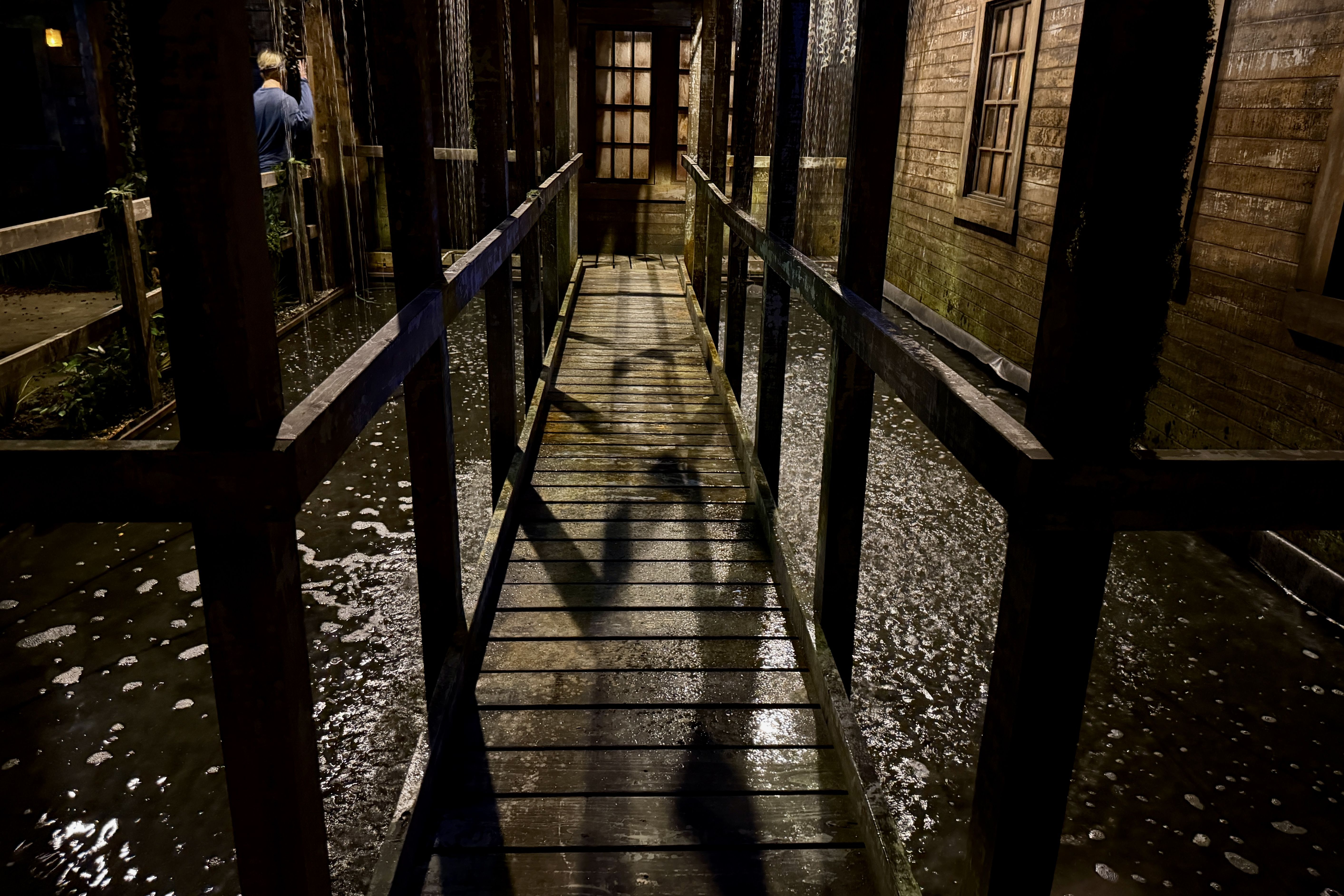 Dimly lit wooden walkway over a narrow canal, with vertical posts. A person in a blue shirt stands left, touching a post; water below, foam, mossy walls, and a windowed building to the right.