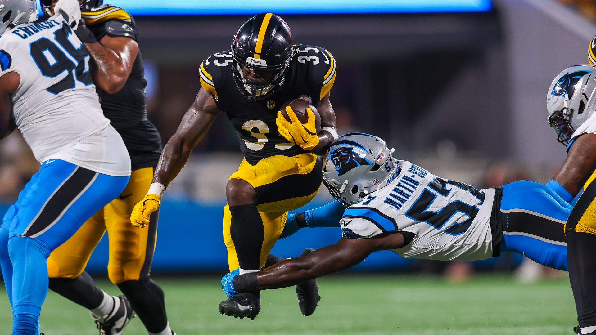 Football player in black and yellow Steelers uniform dodges tackle by player in white and blue Carolina Panthers uniform on green field during game.