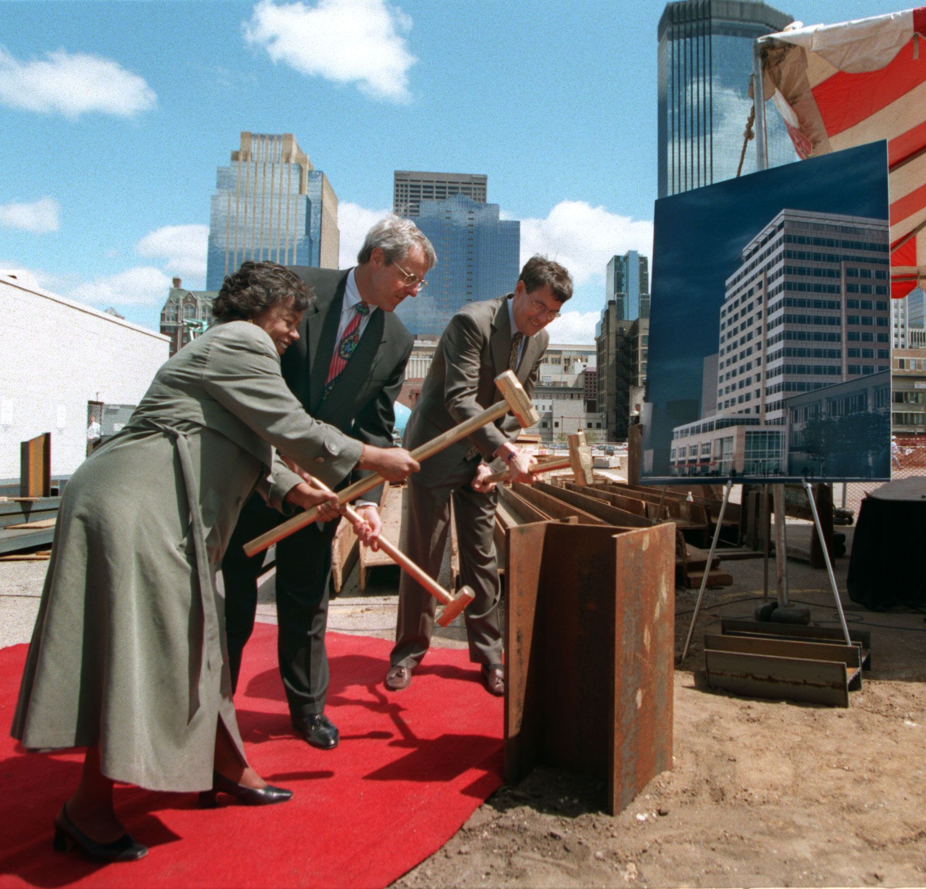 Three suited professionals hammer wooden beams at a construction site, standing on a red carpet with city skyscrapers in the background and a large building poster on the right.