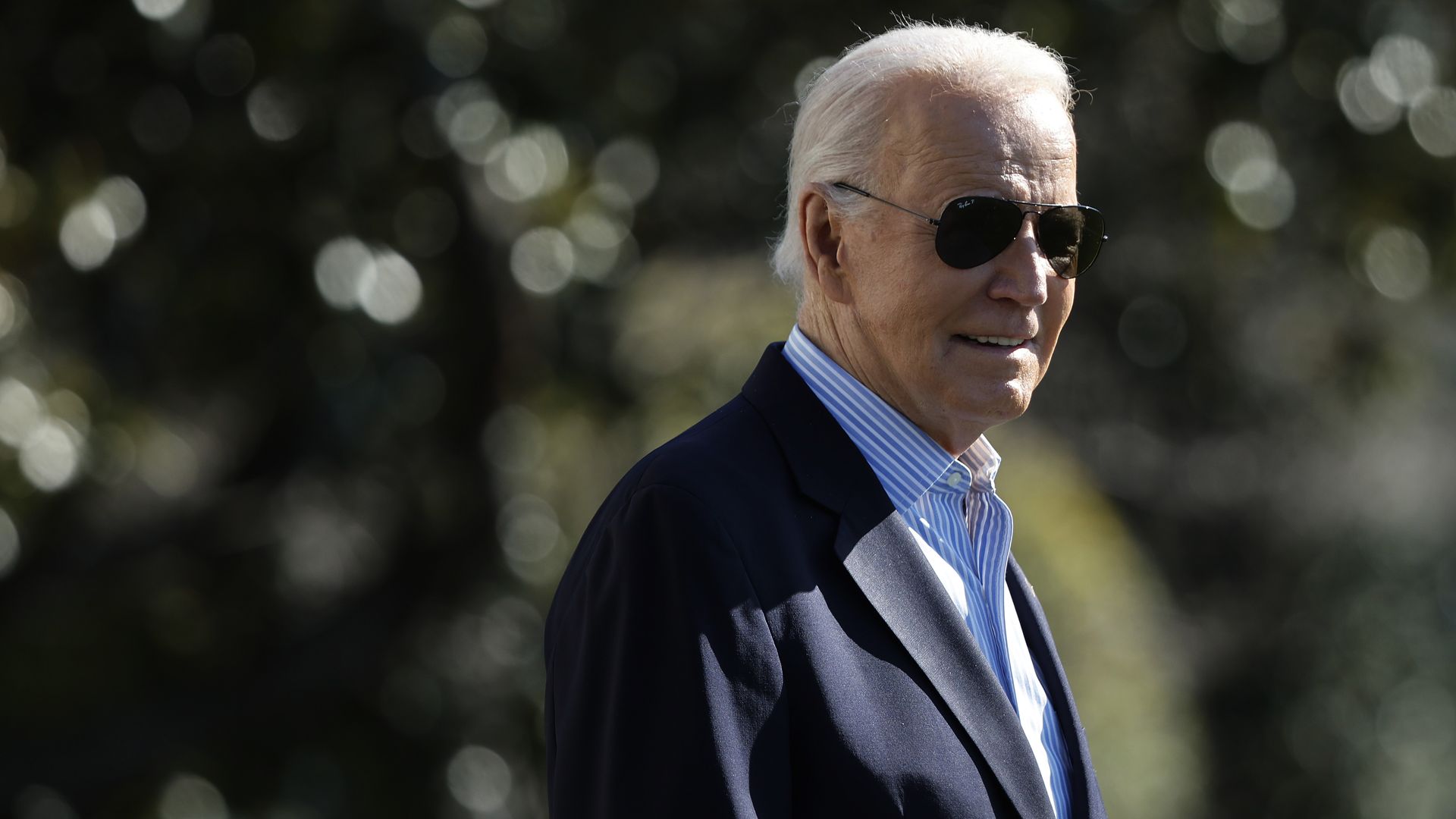 Joe Biden, wearing a dark suit, blue shirt and sunglasses, walks outdoors. 