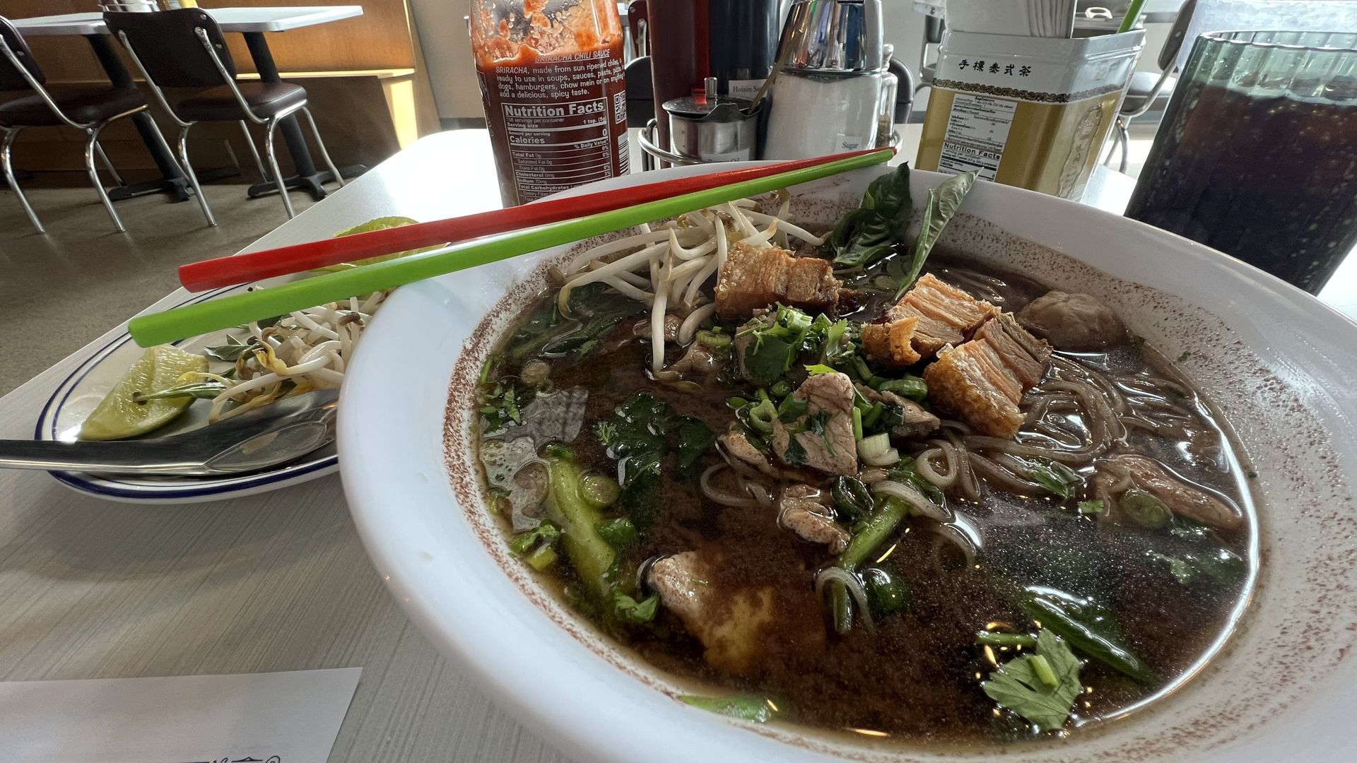 A large bowl of soup with a dark brown broth, pork belly, mint leaves, sprouts, various herbs and rice noodles.