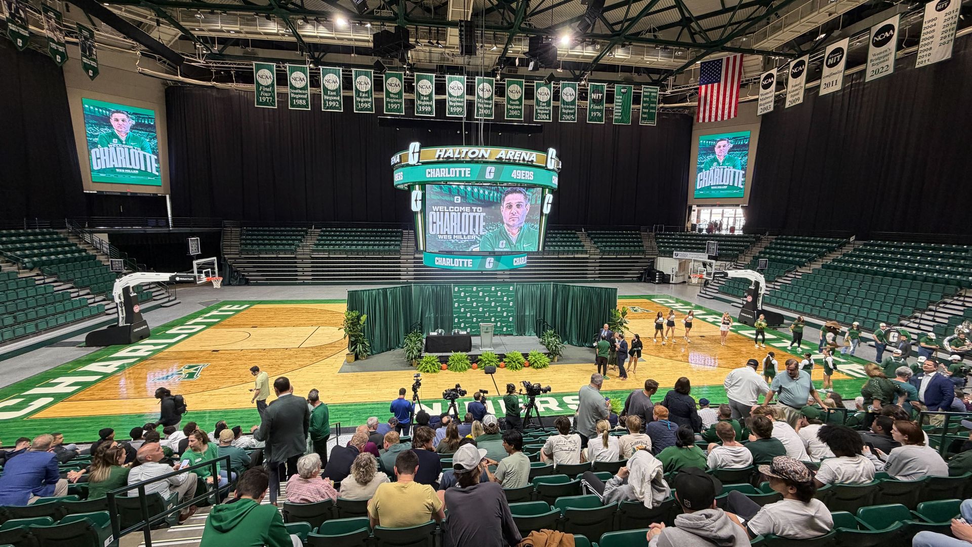 Indoor basketball arena with green seats and banners, a circular scoreboard above a central stage with green drapes, and a crowd gathered on the court for an event.