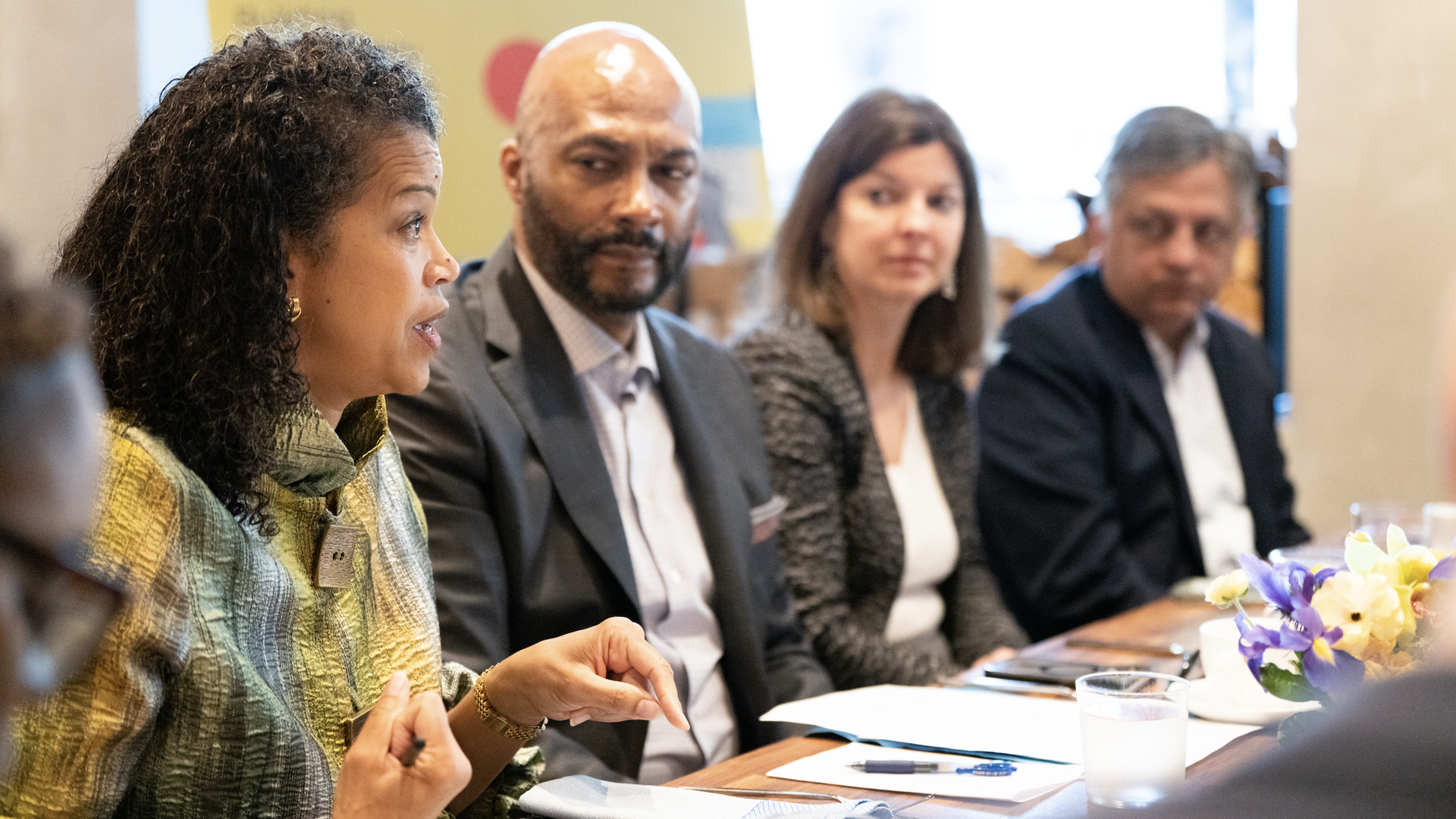 Four professionals sit at a table in a meeting. A woman in a green-yellow patterned top speaks; the others, including a bald man in a suit and a woman in a gray blazer, listen. Flowers on the table.