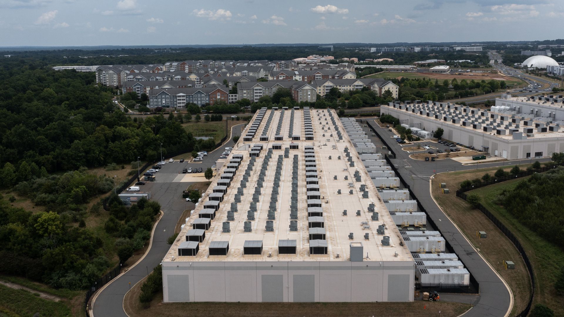 Aerial view of a large data center with rows of HVAC units on a flat roof, surrounded by a residential area, curving roads, and green trees, with a white dome building in the distance under a cloudy sky.