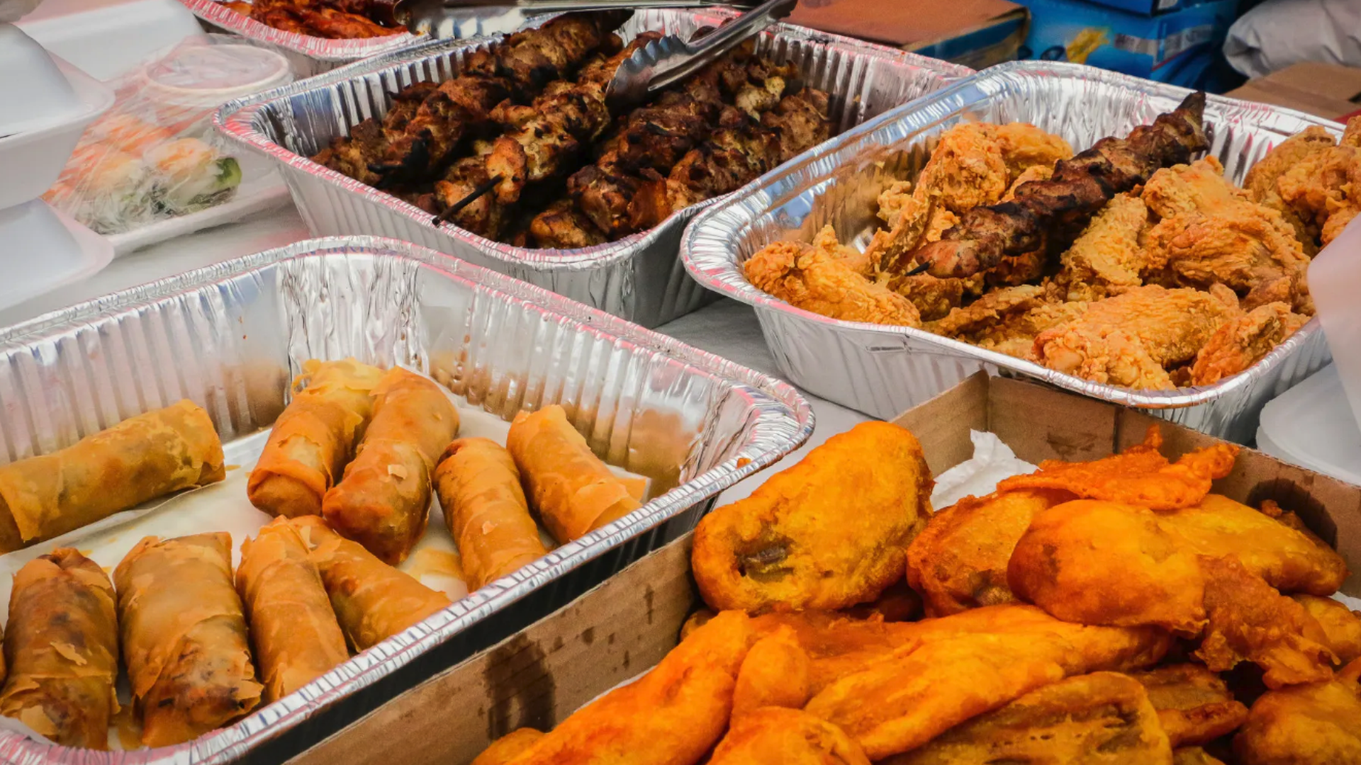 Trays of assorted fried and grilled foods including spring rolls, fried chicken, battered fried fish, and skewered grilled meat, arranged on a table for serving.
