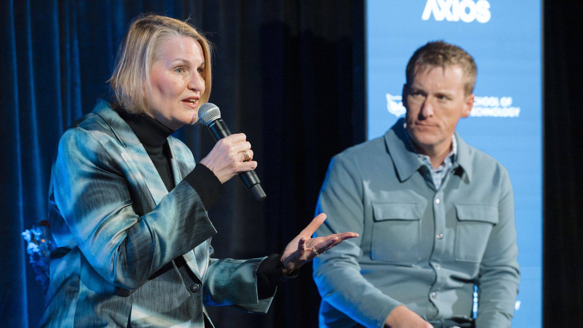 Two panelists on stage: a woman in a blue blazer speaks into a handheld microphone, while a man in a gray jacket listens, against a blue backdrop with a white logo.