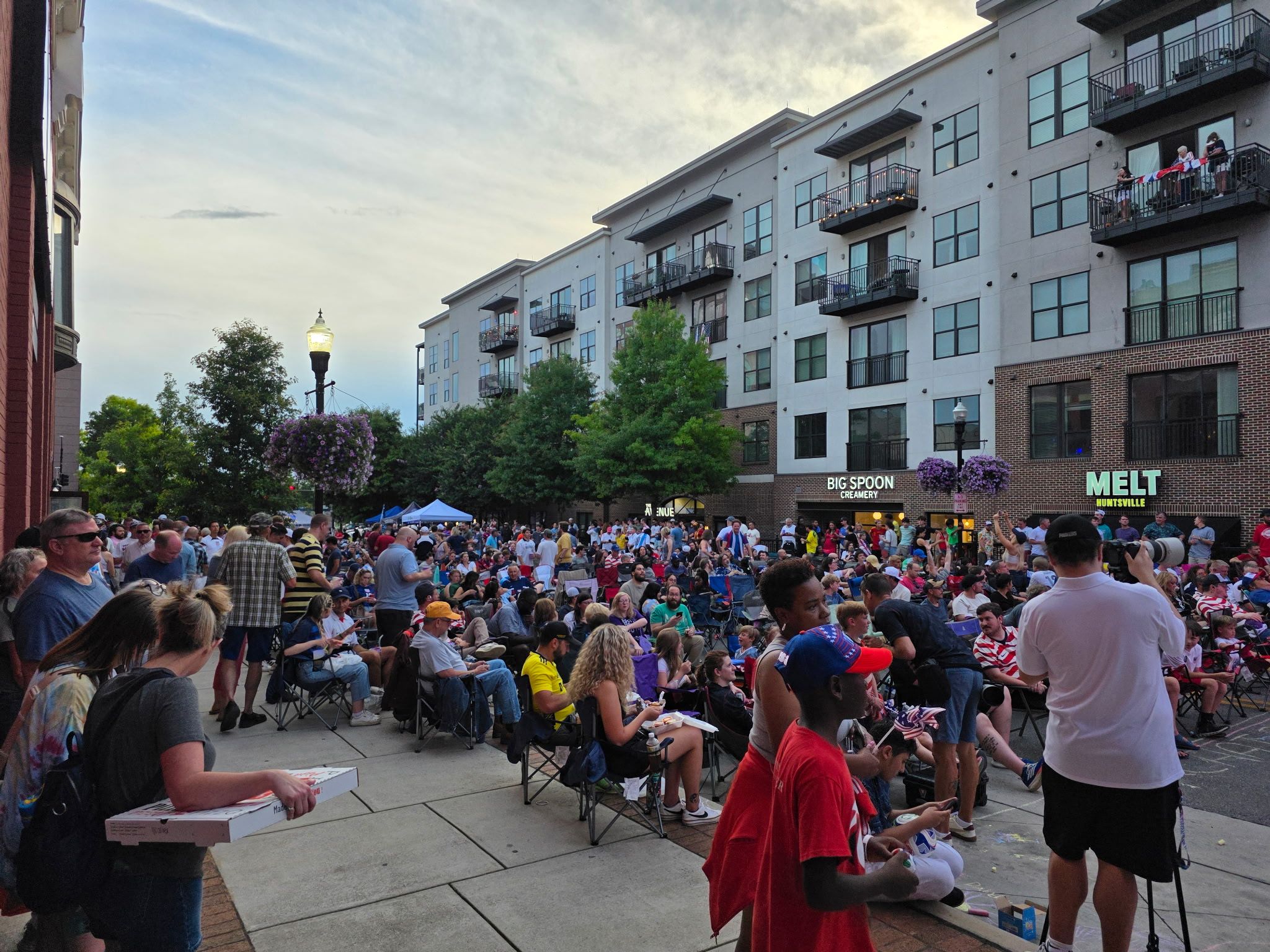 Crowd seated in folding chairs at a street festival, in front of a modern multi-story building with balconies. Storefront signs read "BIG SPOON CREAMERY" and "MELT Huntsville".
