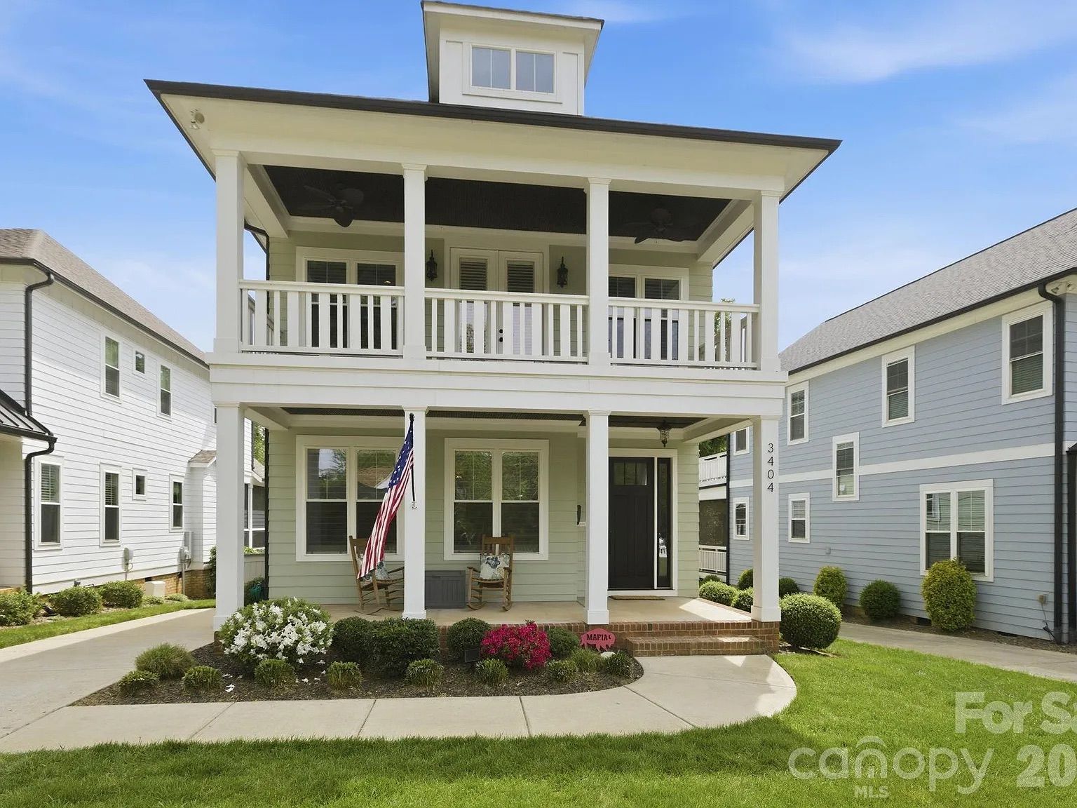Two-story gray house with a white double-deck porch and columns, an American flag on the left column, and a small front garden with white flowers; neighboring pastel houses on each side.