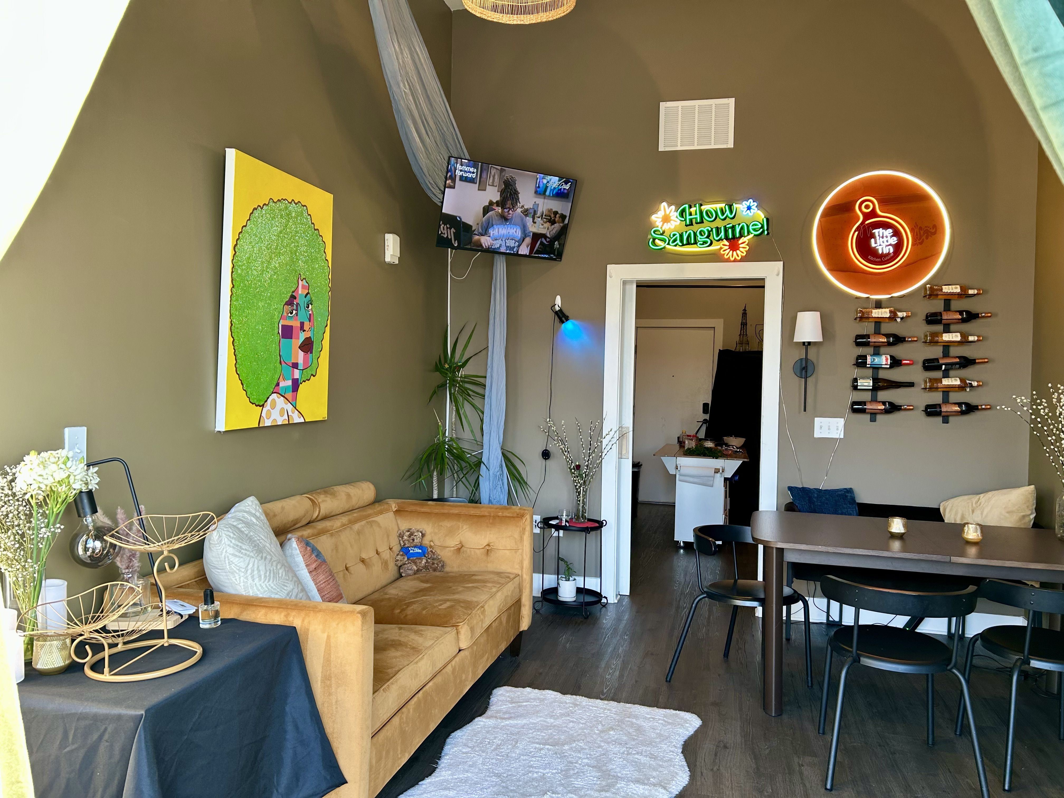 Cozy cafe interior with tan velvet couch, colorful abstract portrait with green hair on olive wall, neon signs reading "How Sanguine!" and "The Little Tin," wine bottle wall display, and dining table.