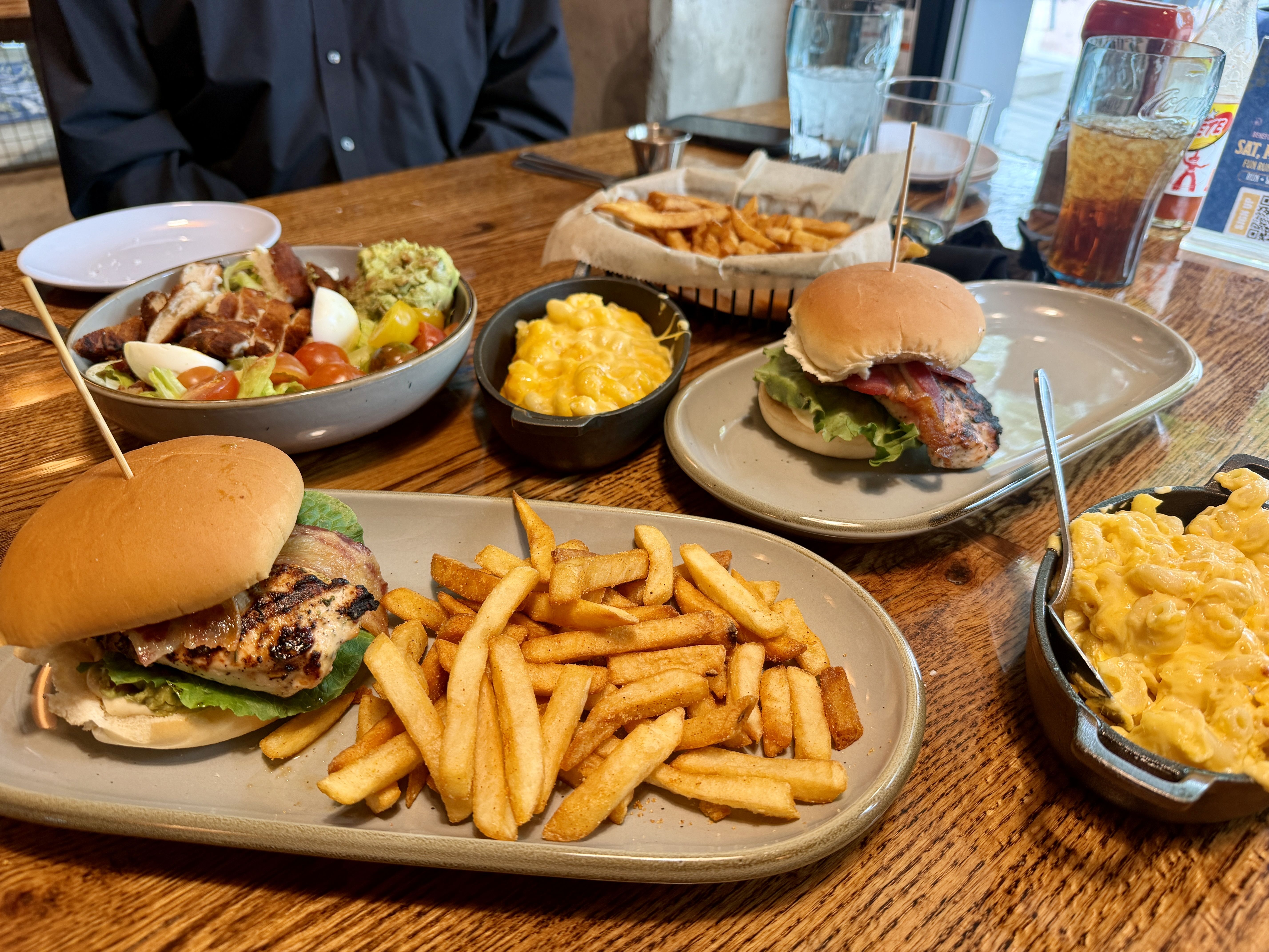Table with two burgers and fries, a skillet of macaroni and cheese, and a salad with greens, tomatoes, steak slices, eggs, and avocado; a basket of chips; person in a dark shirt in background.