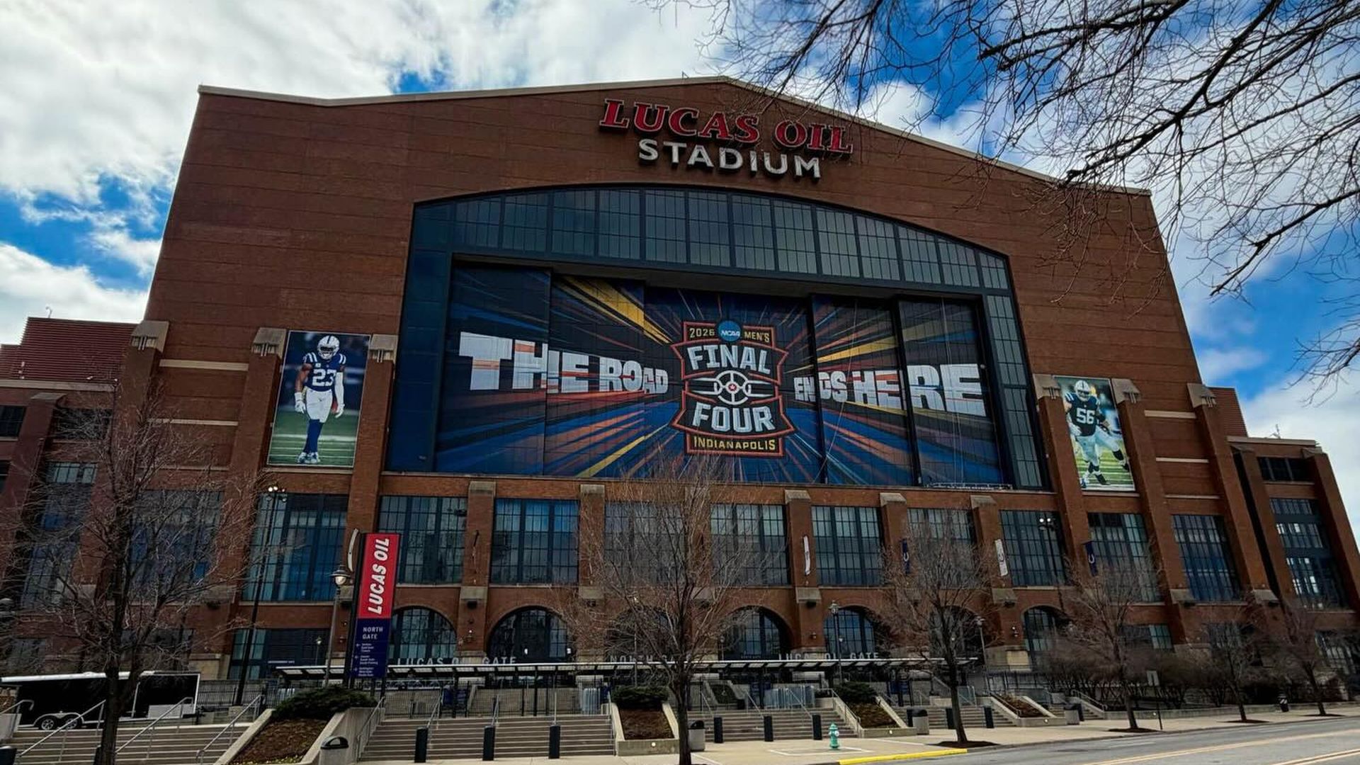 Front view of Lucas Oil Stadium, a red-brick arena with a large blue banner reading \"Final Four\"; bare trees, an empty street, and a blue sky with clouds.