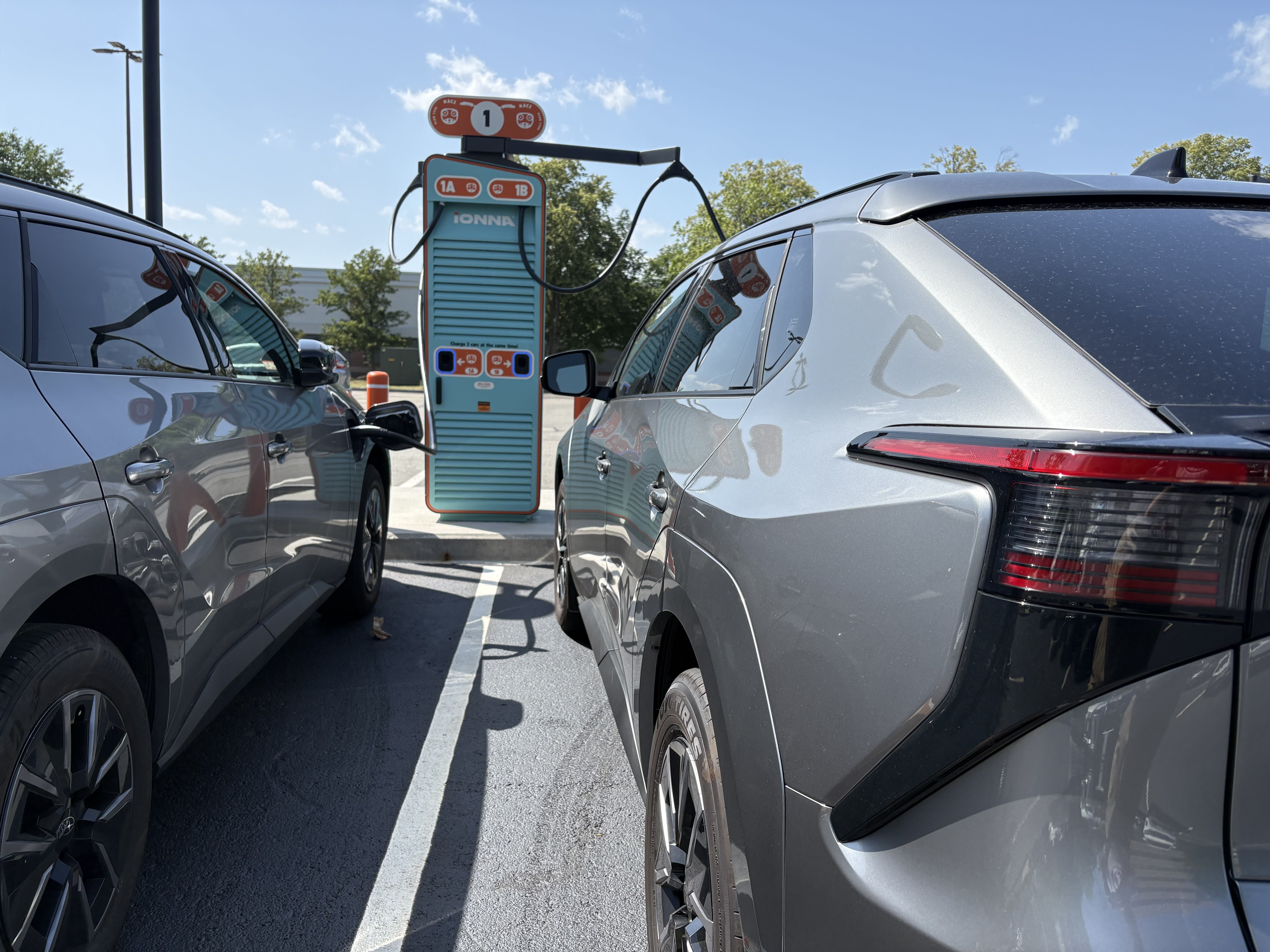 Two silver SUVs flank a teal retro-style Ionna EV charging station outdoors; cables connect to the cars as a bright blue sky and trees loom in the background.