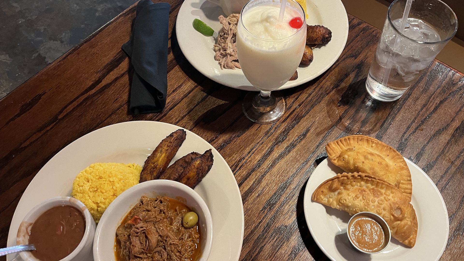 Top-down view of a wooden table with plates of food: yellow rice and shredded beef plate with corn and plantains; a frothy milkshake with a cherry; coffee mug; glass of ice water; and pastries with sauces.