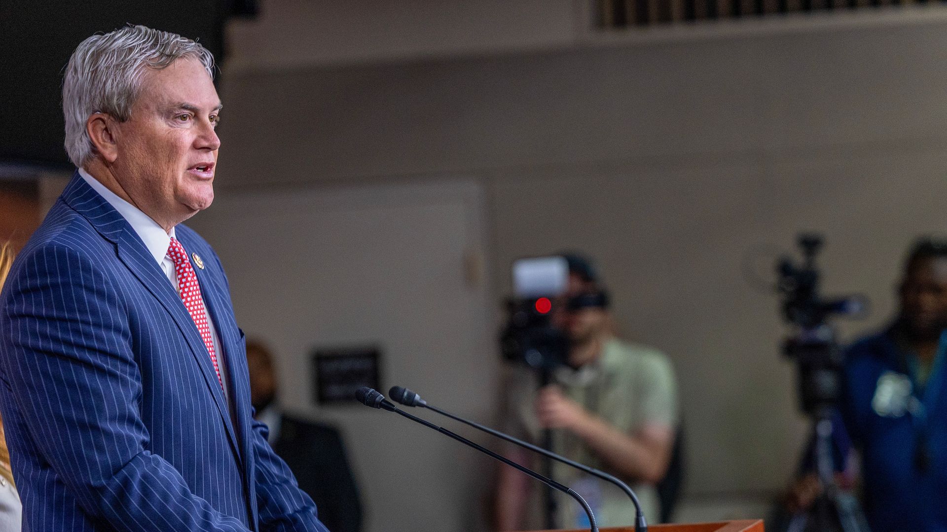 James Comer wears a striped blue suit, a collared shirt and a red tie while he speaks into microphones from behind a podium.
