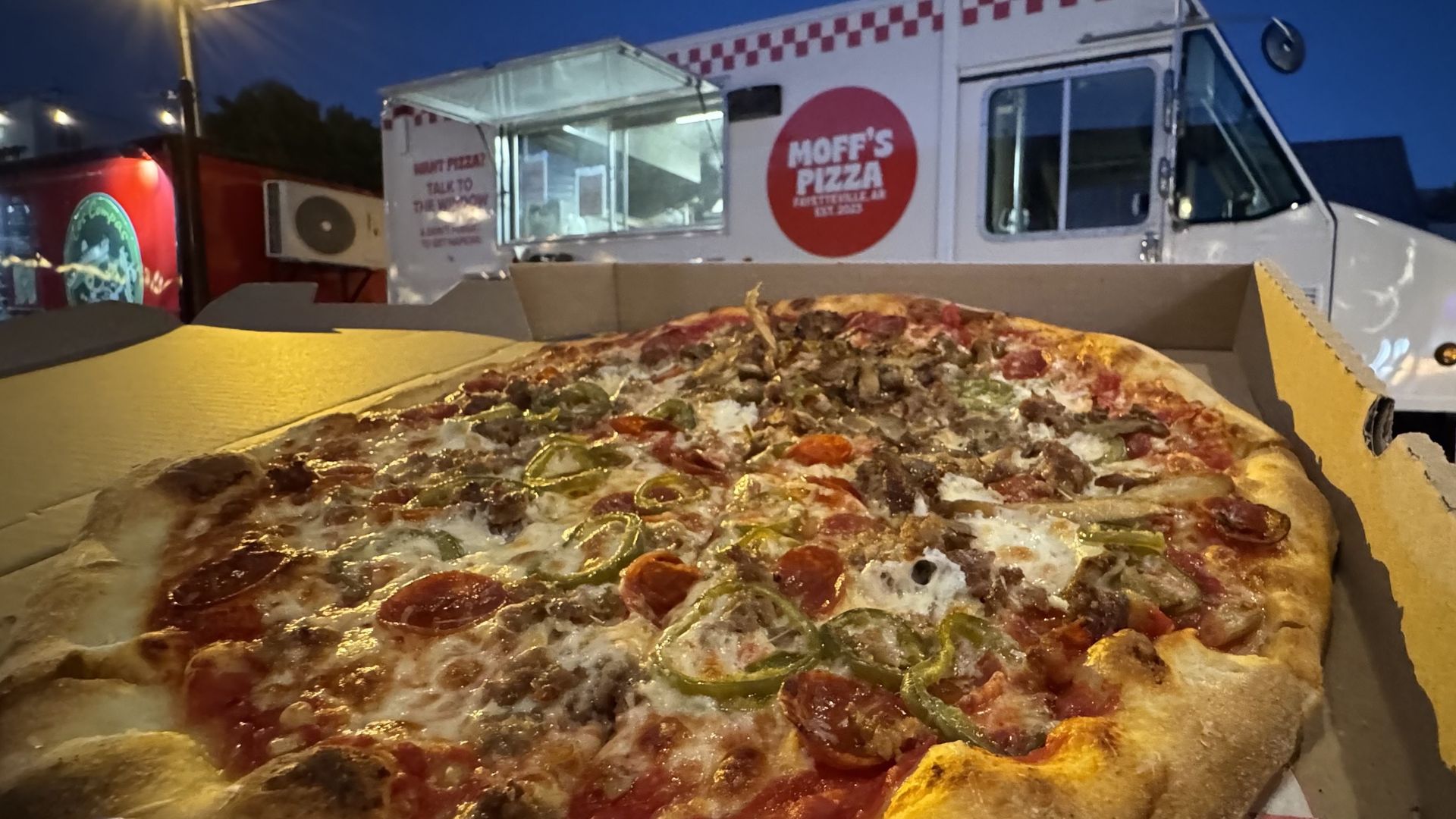 A photo of a pizza in a box with a food truck in the background.