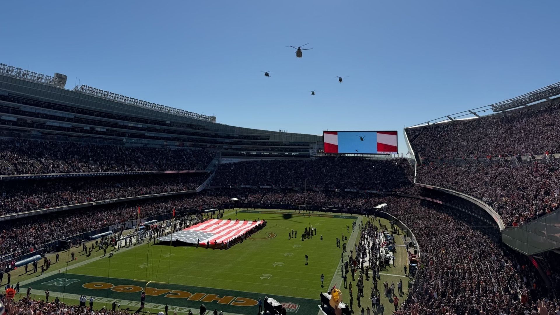 Wide view of a packed football stadium on a sunny day. Four helicopters hover in the clear blue sky. A large screen shows a red, white, and blue banner; the field features a red-and-white striped display.