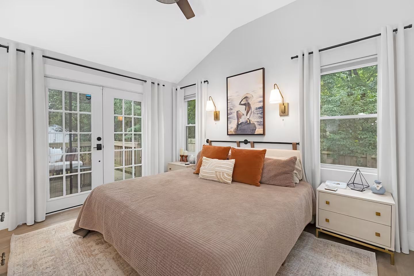 Bright bedroom with vaulted ceiling, large bed with brown quilt and rust-colored pillows, white curtains framing French doors and windows showing green trees outside, wall sconces, and abstract art.