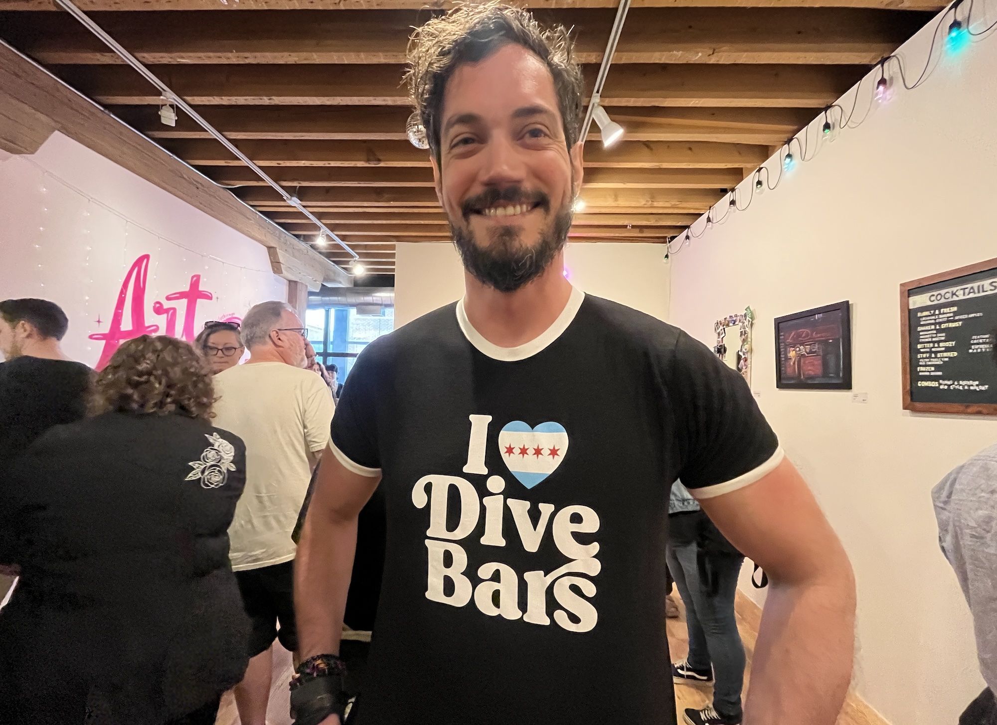 Smiling man with a beard wearing a black t-shirt that reads I Love Dive Bars, featuring a heart icon; stands in a casual space with wood-beam ceiling, string lights, and people in the background.