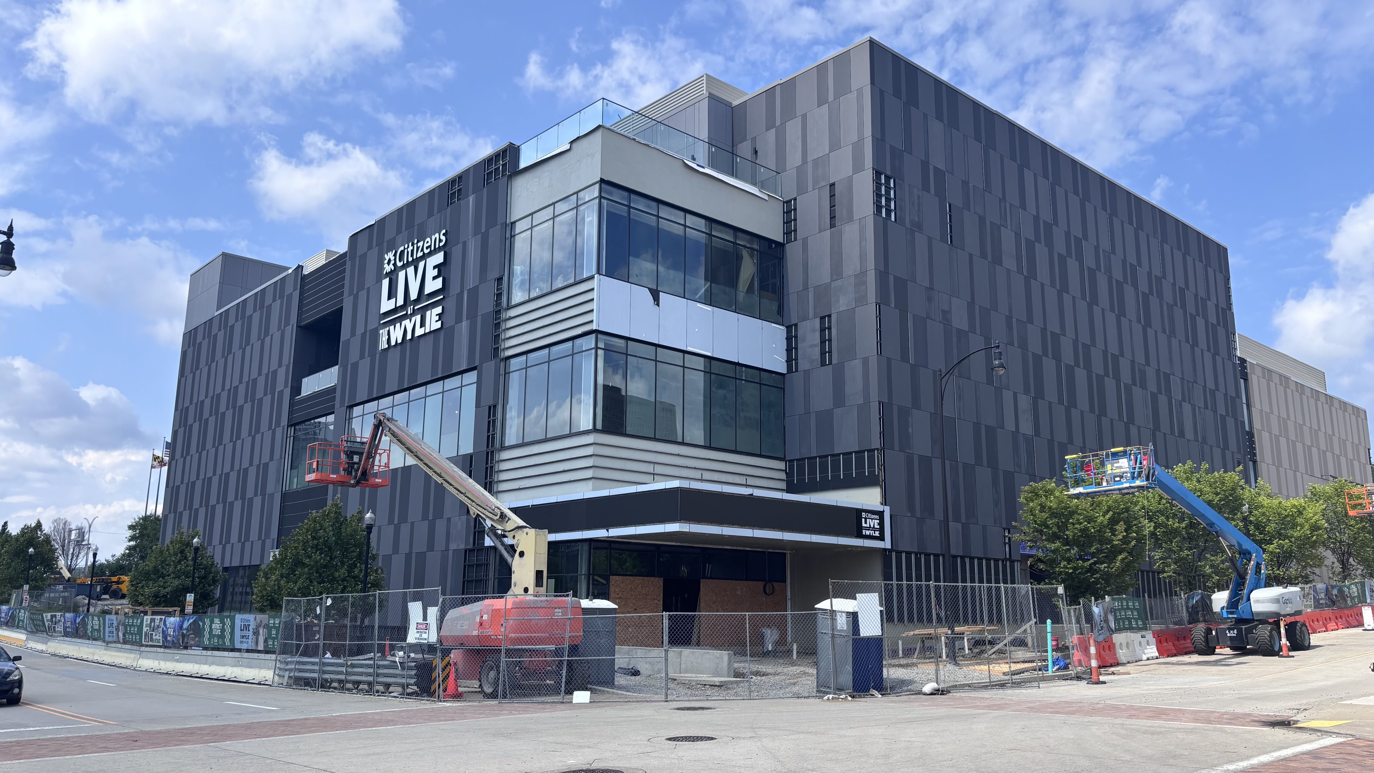 Gray, glass-clad building under construction with a sign reading "Citizens LIVE at THE WYLIE" on the facade; construction equipment and fencing in the foreground under a blue sky.