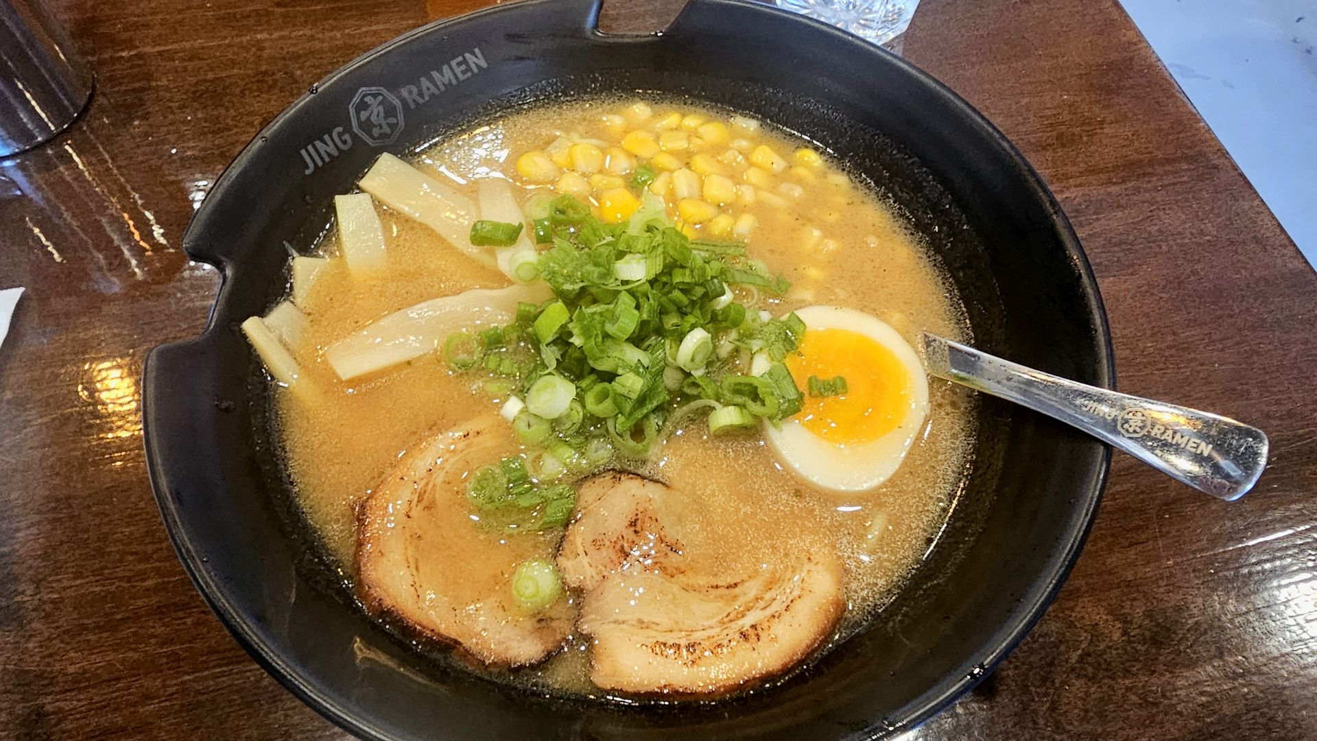 Top-down view of a black ramen bowl on a wooden table, filled with creamy broth, corn, green onions, bamboo shoots, a halved soft-boiled egg, and pork chashu; a spoon rests in the bowl.
