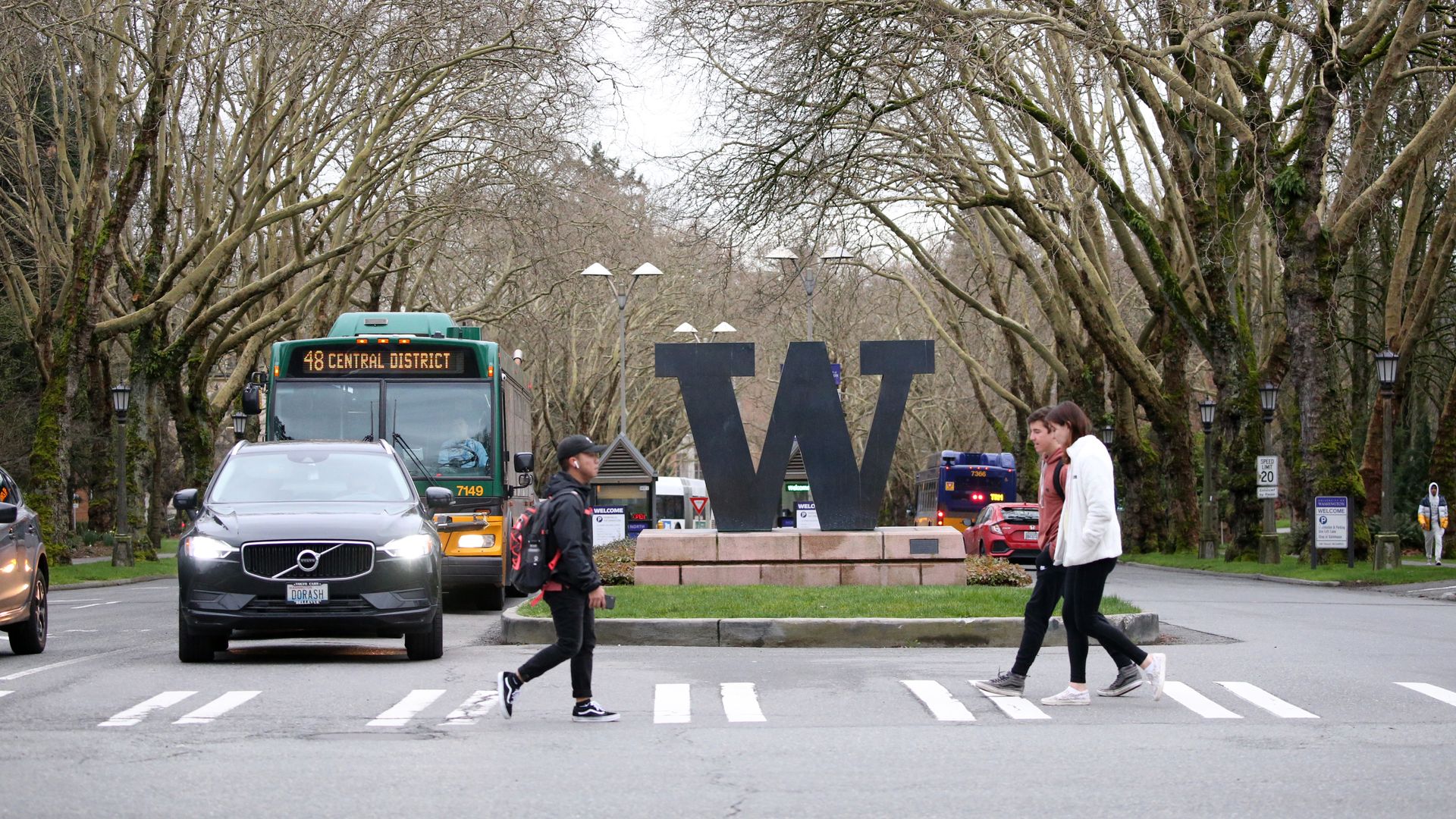 People walk along a crosswalk in front of the "W" sign at the entry of the University of Washington, flanked by trees and traffic.