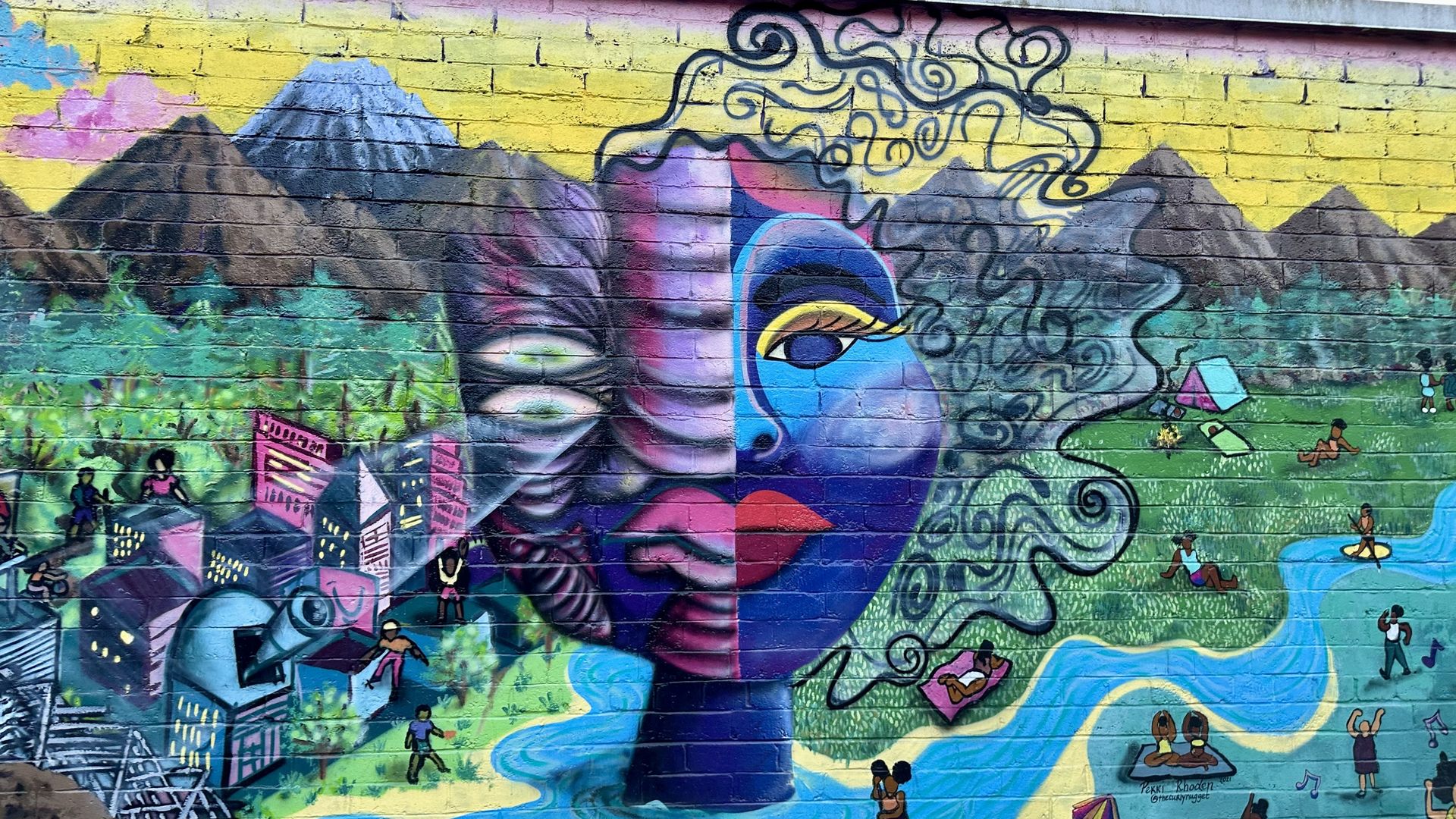 A colorful mural with mountains, a golden sky, river and a person with some alien-like features.