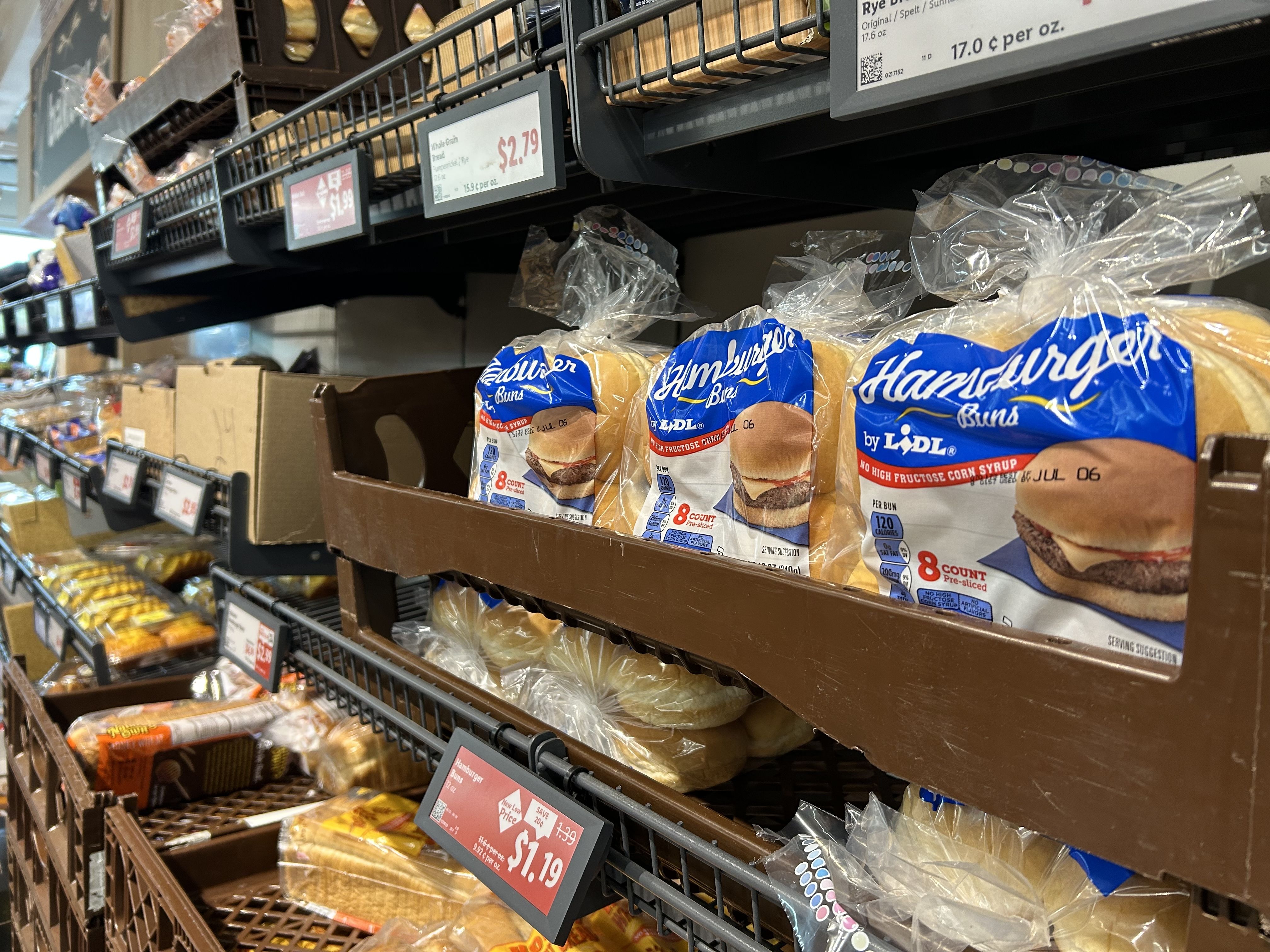 Store bakery aisle with brown metal shelves and bags of blue-labeled Hamburger Buns by Lidl, visible price signs showing $2.79 and $1.99.