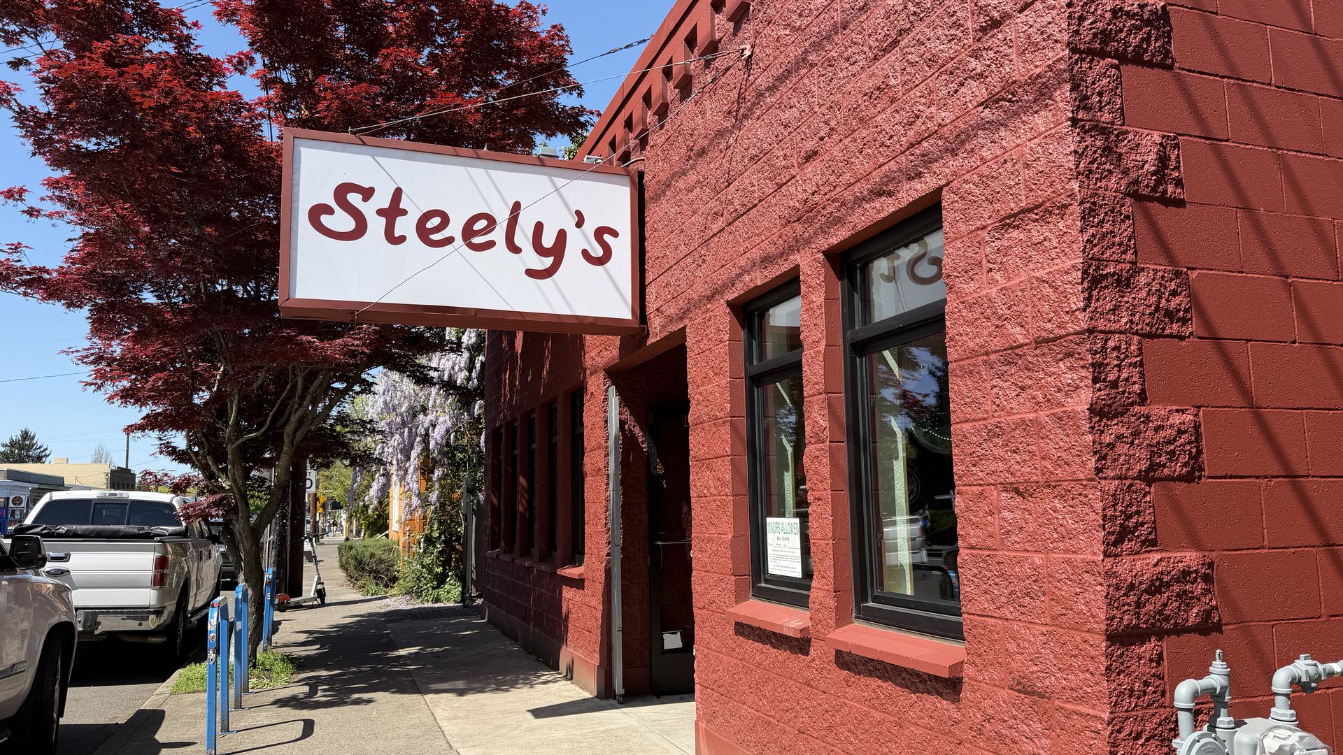 Street view of a red brick building with a white sign reading "Steely's" hanging above a sidewalk. A blue-sky day, a red-leaf tree, parked vehicles, and blue bike racks line the street.
