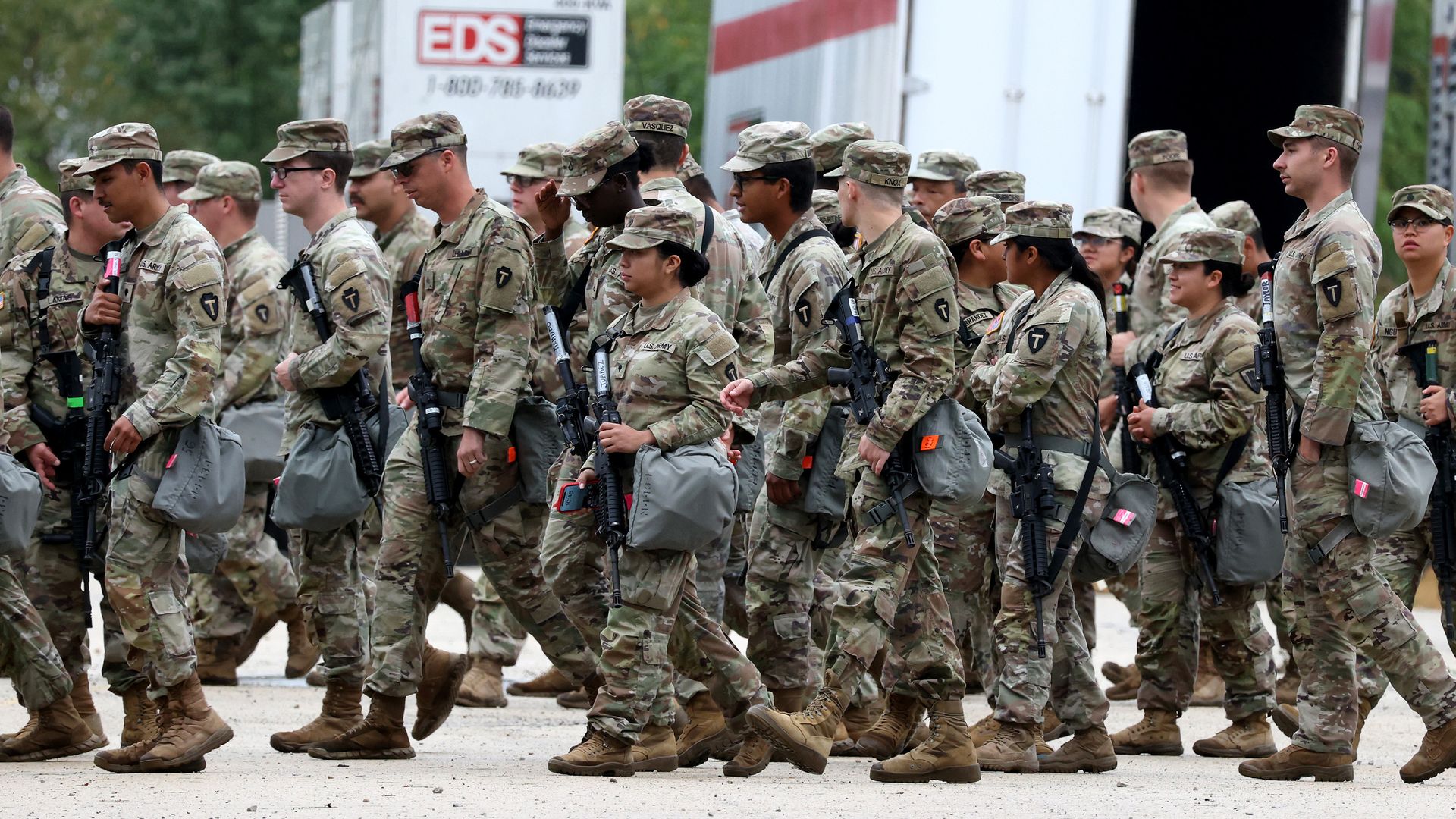 Group of U.S. Army soldiers in camouflage uniforms and caps walking, carrying rifles and gear, with trucks and greenery in the background.