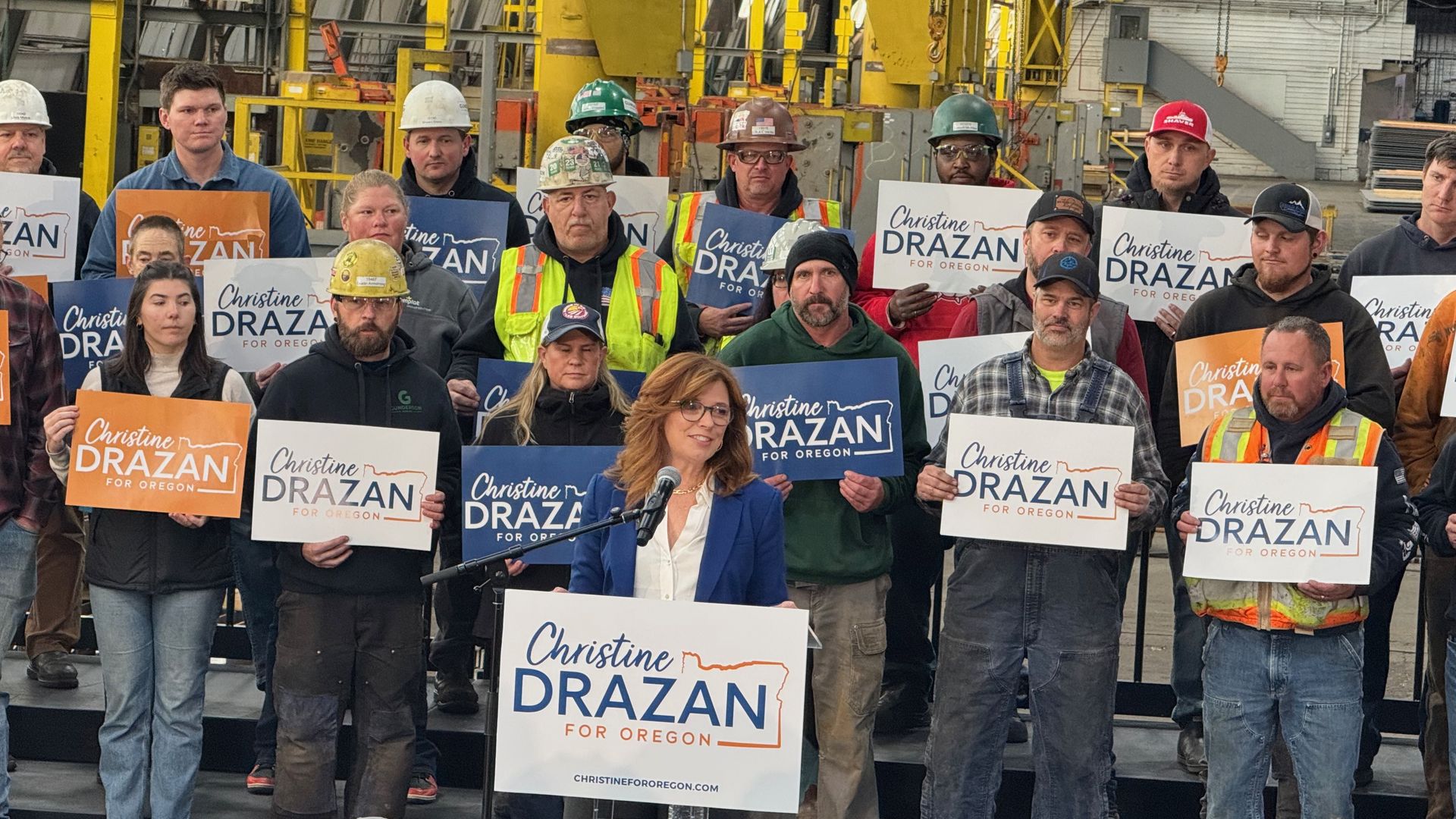 A diverse group of workers in hard hats and safety vests hold signs reading "Christine Drazan for Oregon" at a factory, with a speaker at a podium in the foreground.