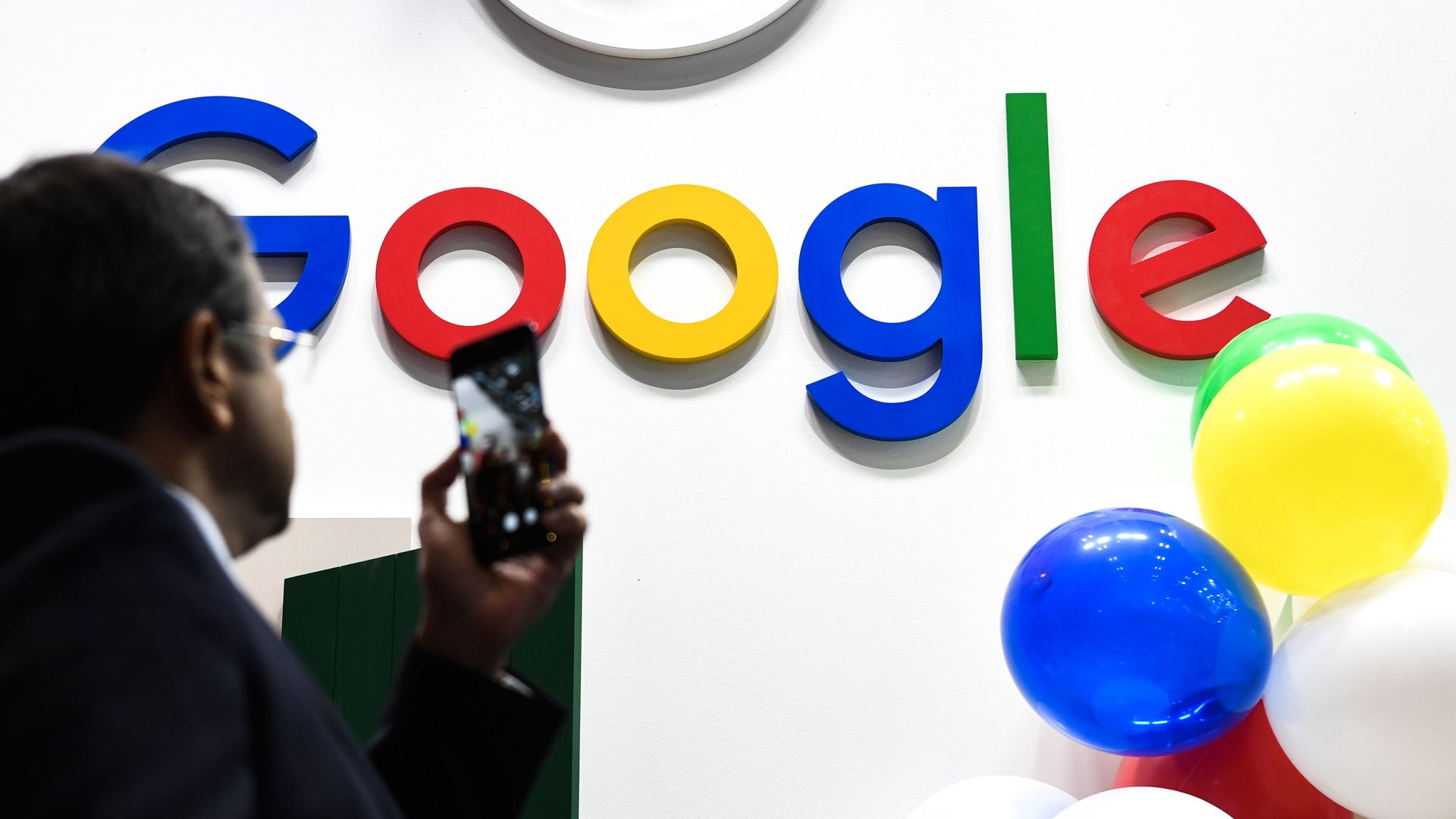 In this image, a man takes a picture of the Google logo as balloons float nearby.