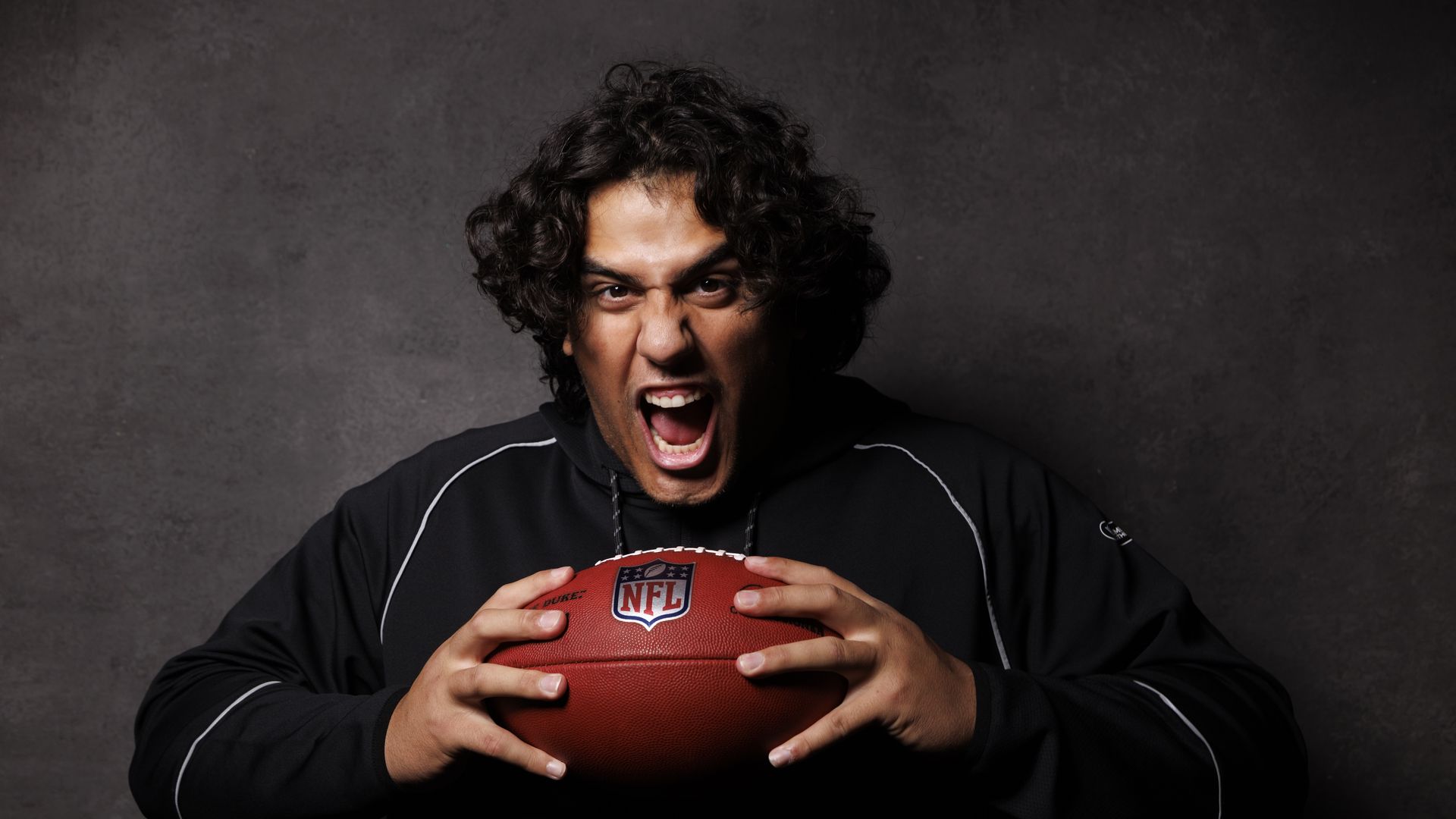 A man with curly dark hair wearing a black jacket grimaces with his mouth open, gripping a red NFL football with both hands against a dark textured background.