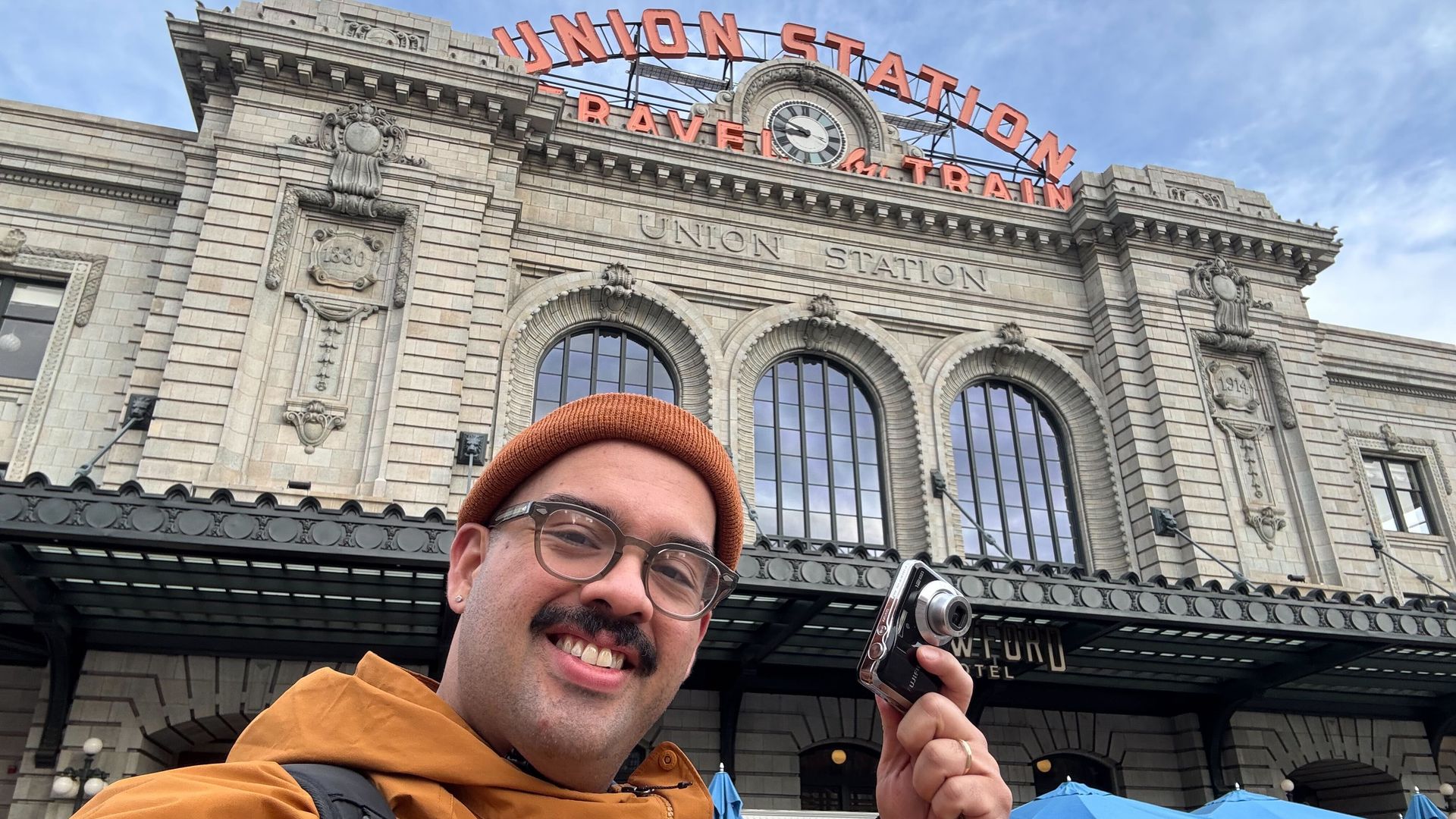 Selfie of a man with a beanie and glasses in a brown jacket, backpack, in front of Denver's Union Station's stone facade with arched windows and a large orange Union Station sign; he holds a small camera.