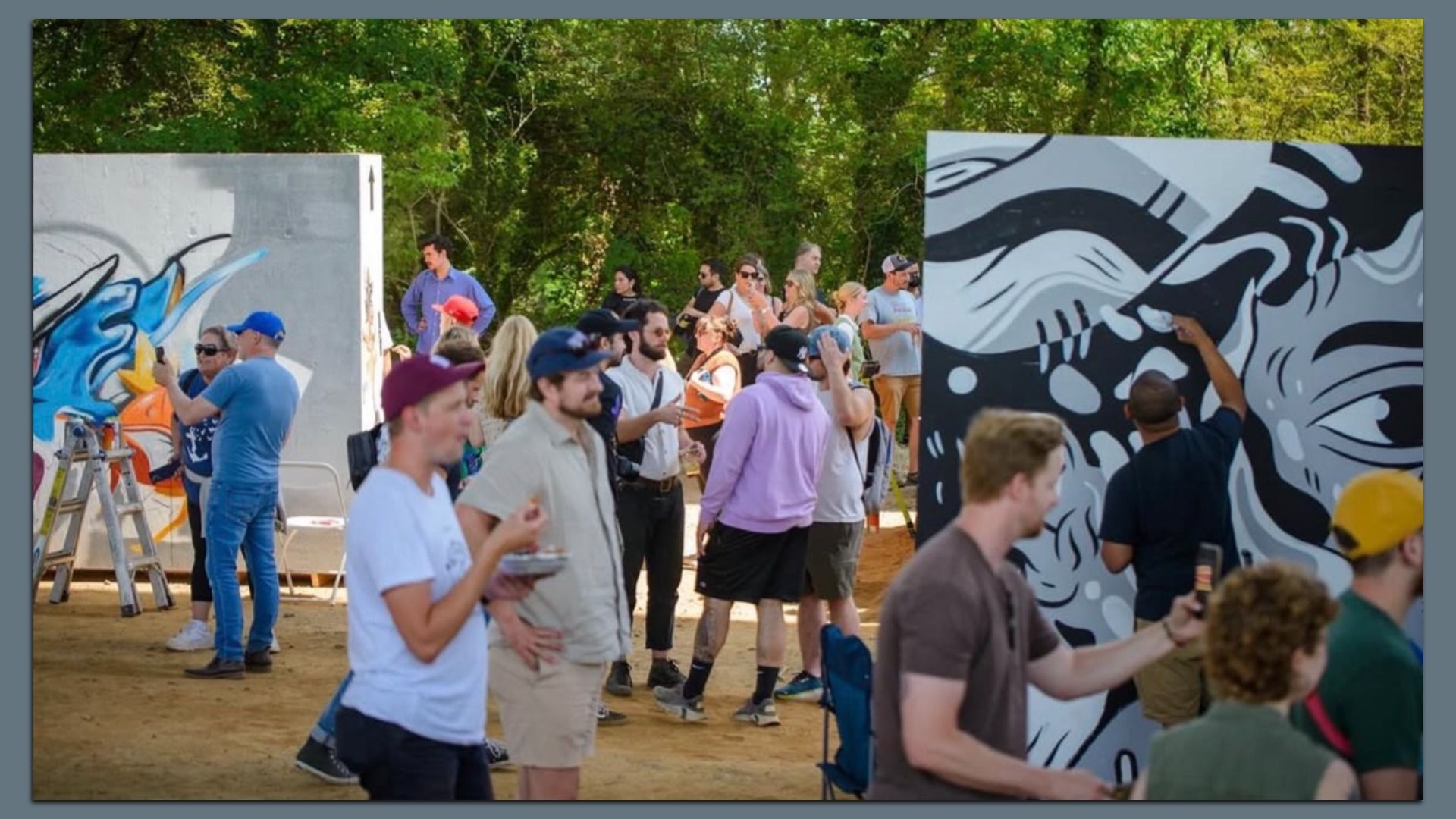 Crowd at an outdoor graffiti art show; artists paint large panels with blue and orange graffiti on the left, and a black-and-white mural on the right. People mingle under trees.