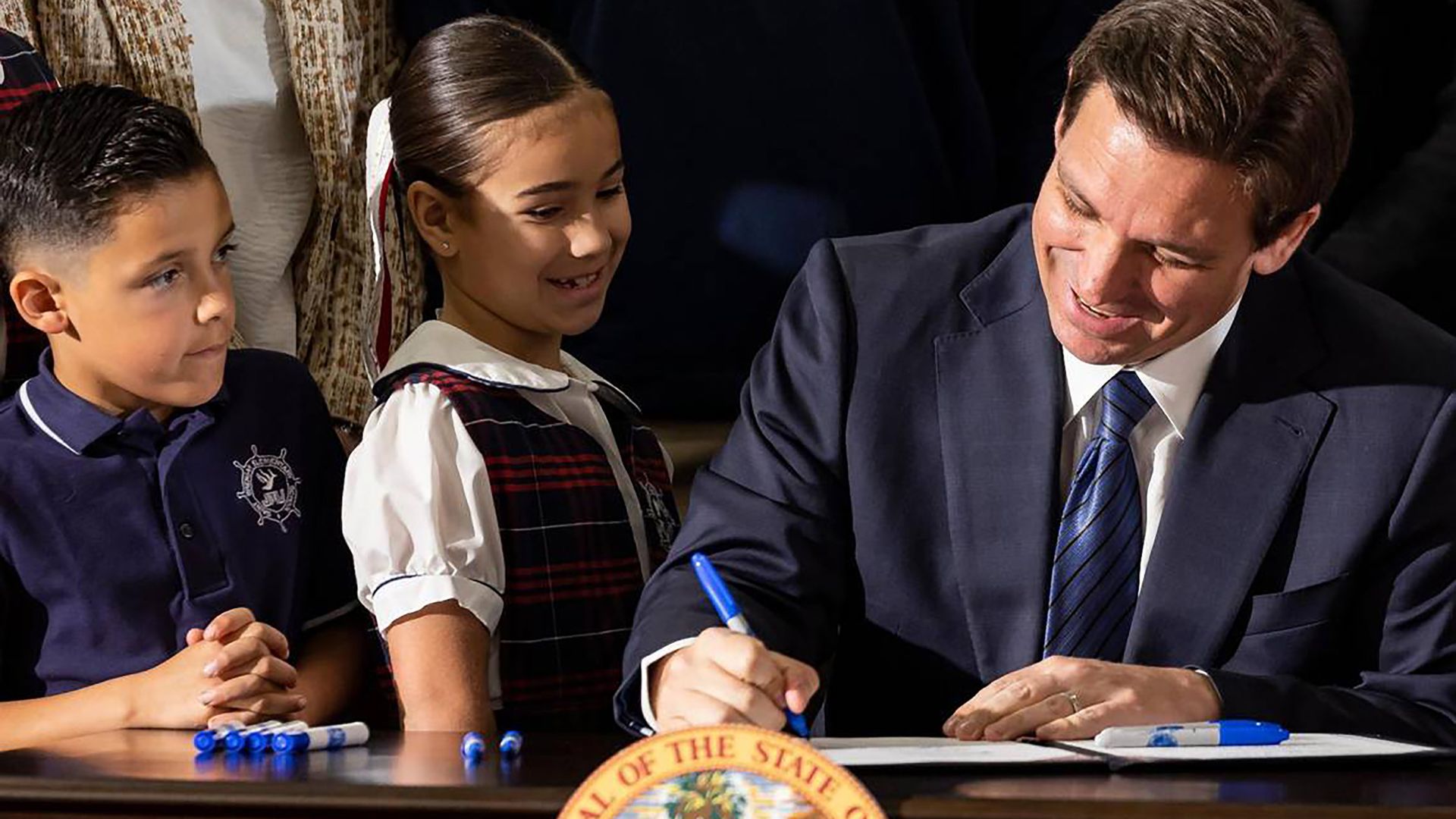 Gov. Ron DeSantis signing HB 1, with two young students next to him.