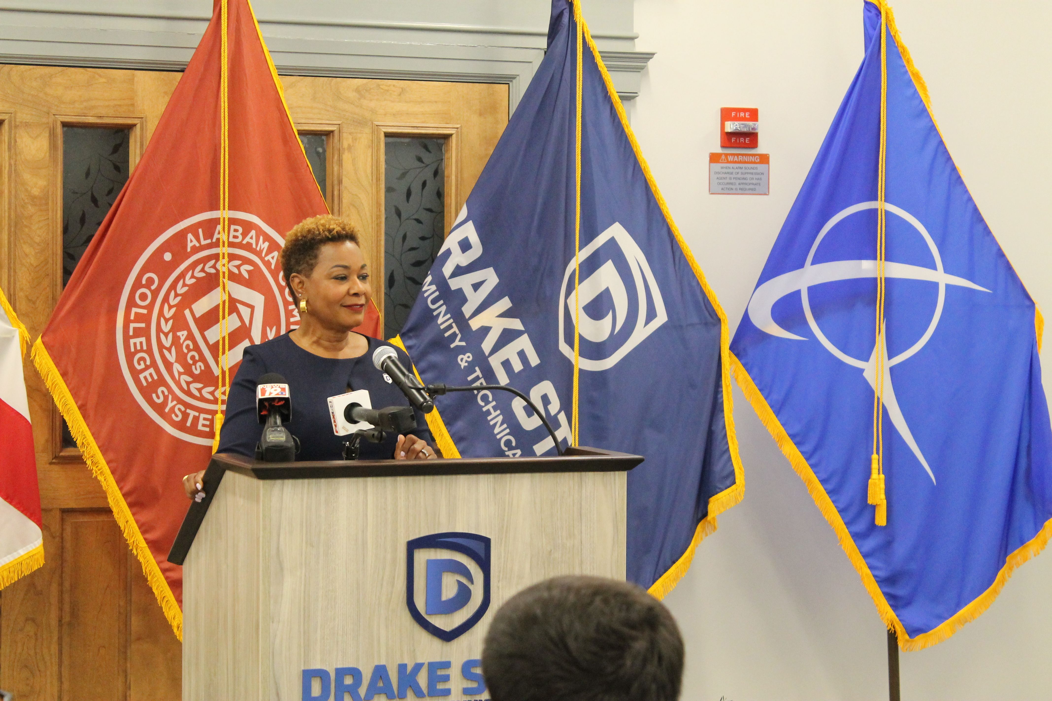 A woman speaks at a podium with microphones, in front of three tall flags with gold fringe: a red Alabama flag, a blue flag with a white logo, and a blue flag with a white circular emblem.