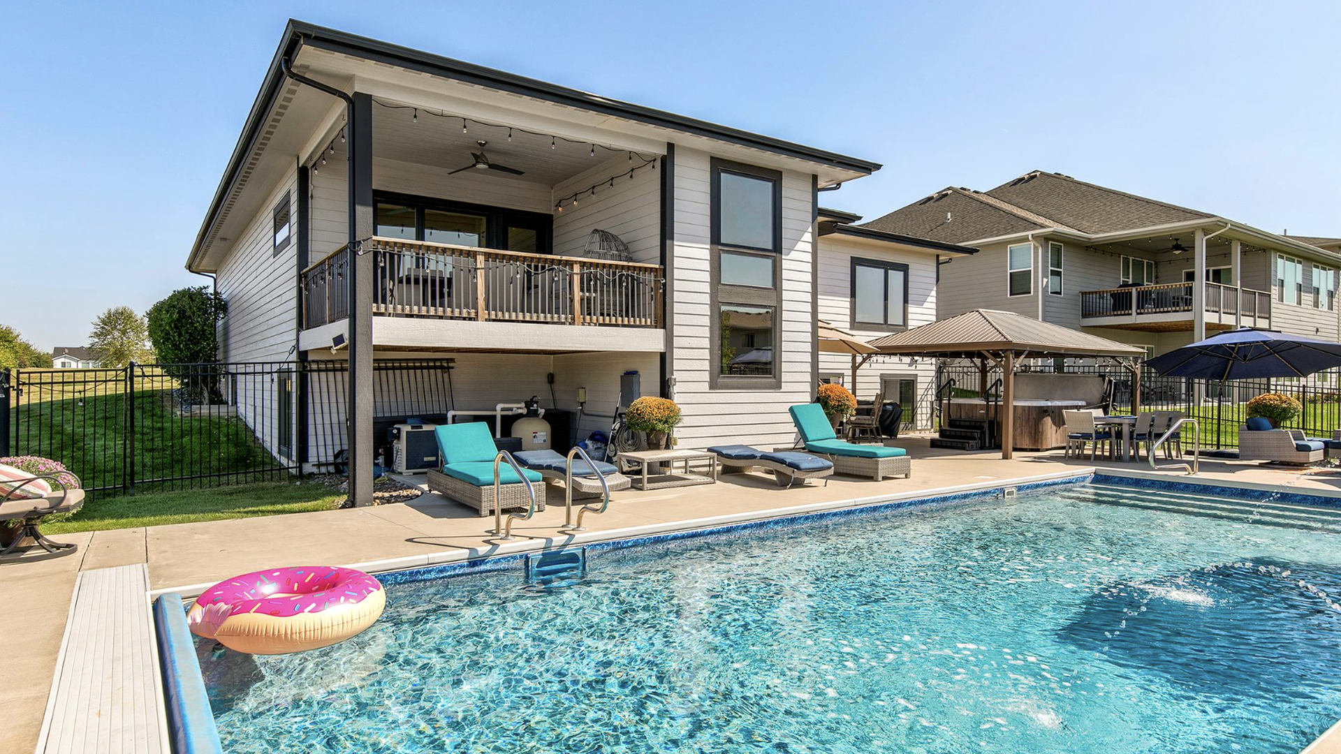 Clear blue pool with a pink donut float next to a modern light gray house and patio featuring turquoise lounge chairs, orange flowers, and a shaded seating area under a sunny blue sky.
