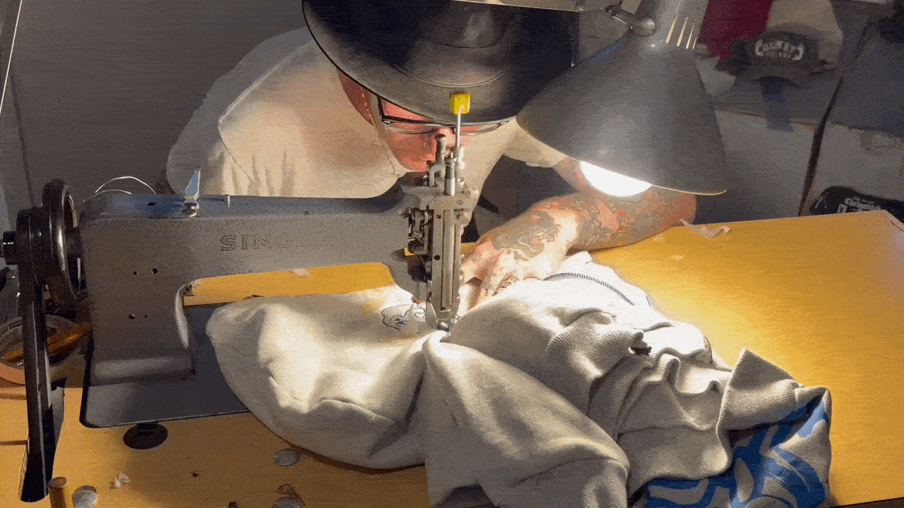 A person wearing a cap and glasses leans over a vintage gray Singer sewing machine, sewing a gray fabric on a yellow worktable under a desk lamp; tattoos visible on their arm.
