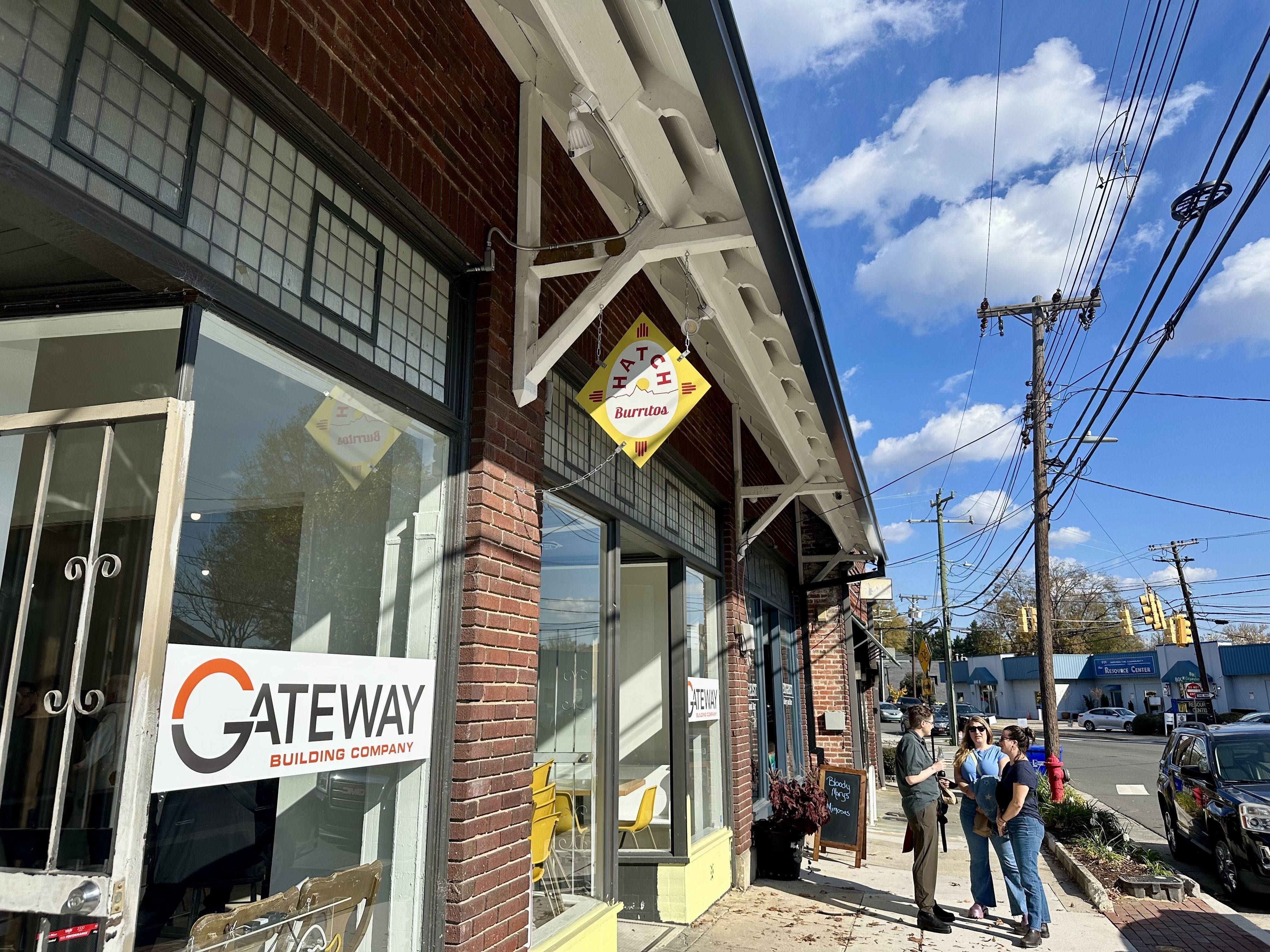 Brick building with signs for "Gateway Building Company" and Hatch Burritos. Three people stand on the sidewalk under a blue sky with clouds and power lines overhead.