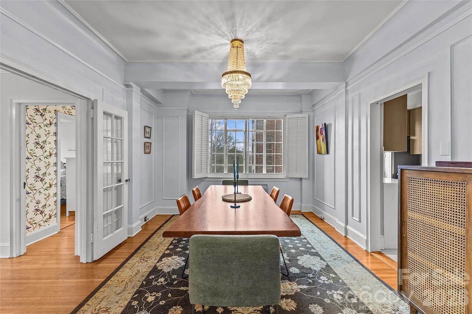 Dining room with lavender walls, wooden table, six chairs, floral carpet, large window with white shutters, crystal chandelier, and floral artwork on the wall.