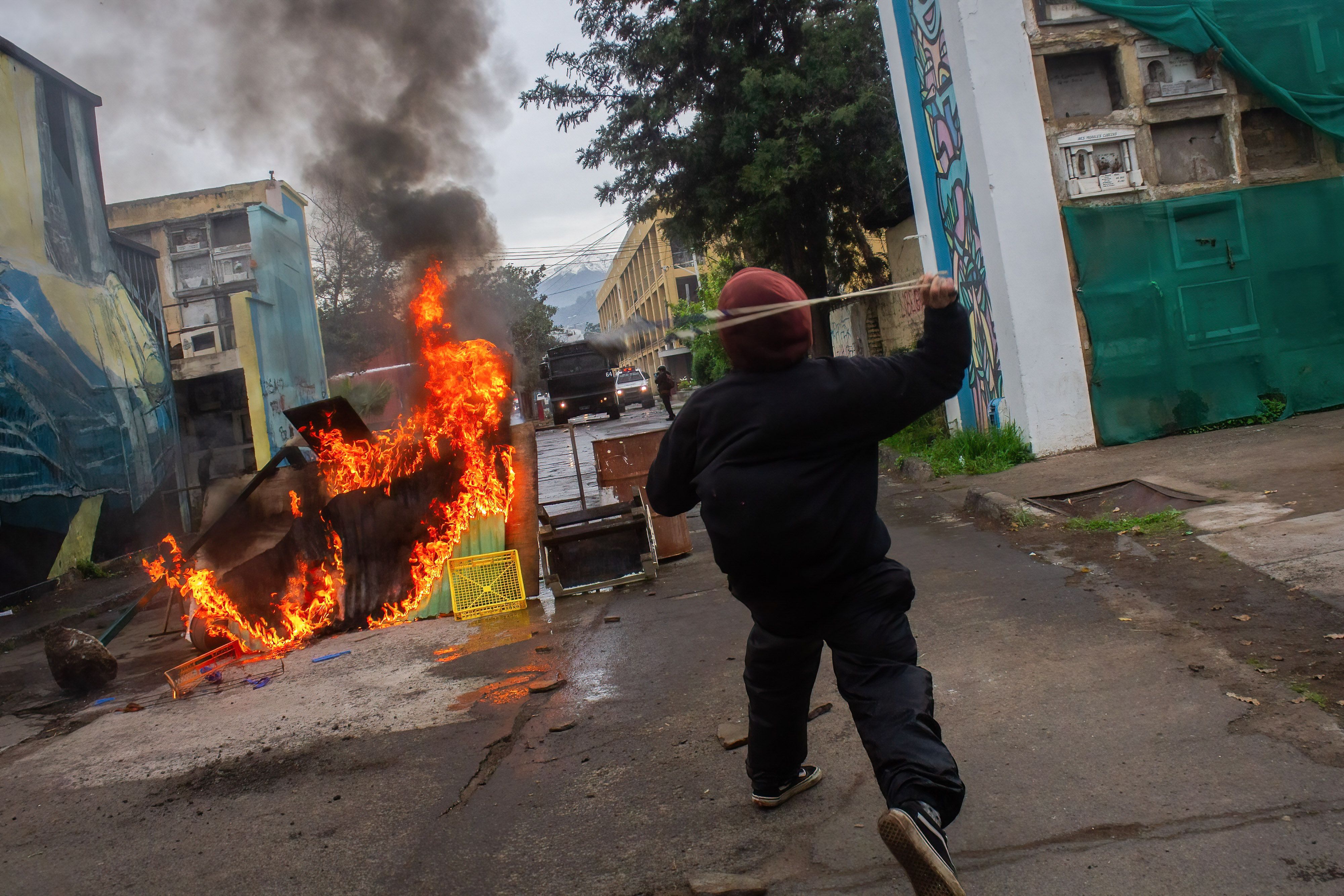A short person, seen only from behind, throws a stick at a flame after a protest in Chile. 