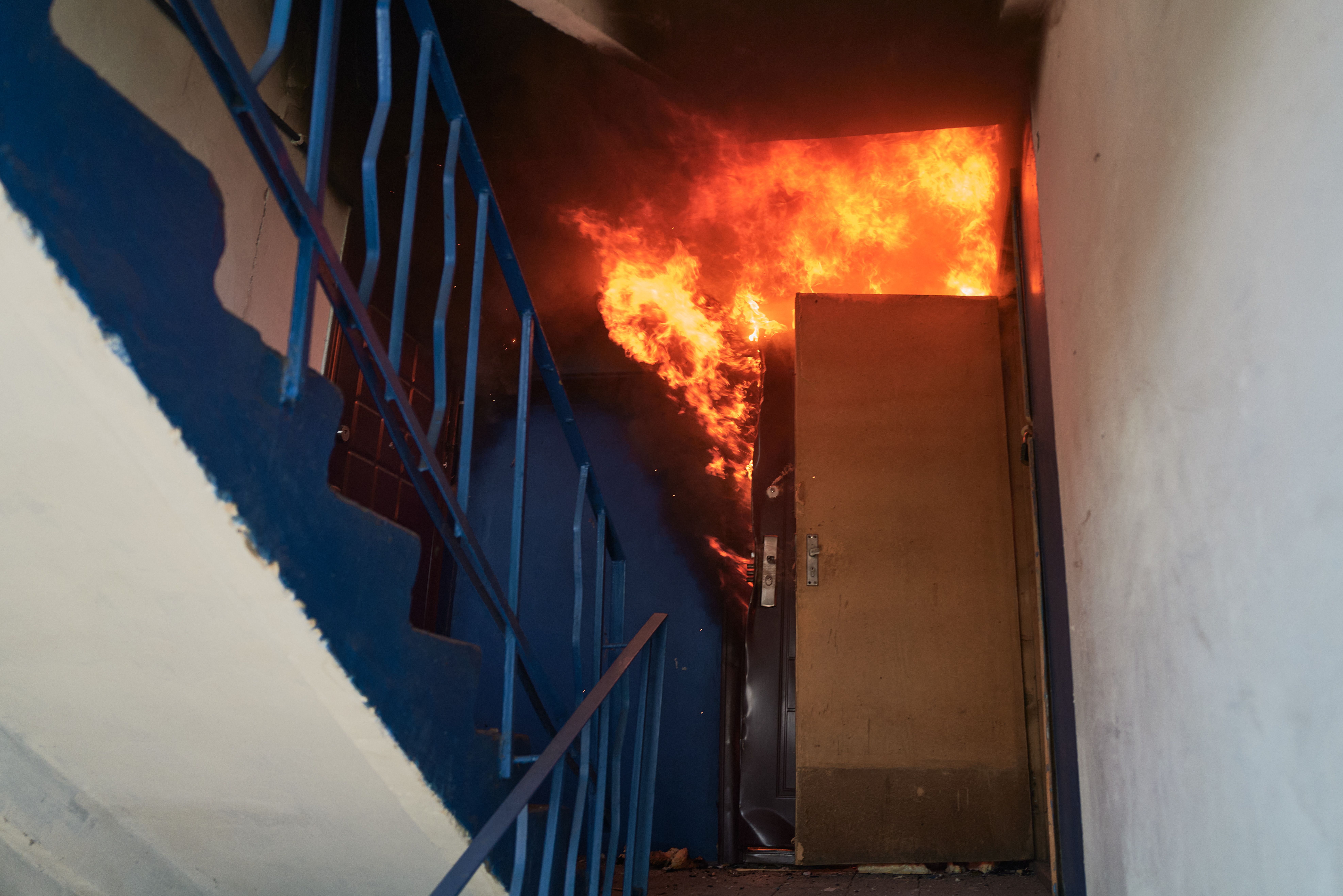 Bright orange flames and smoke engulf a doorway at the top of a blue-painted stairwell inside a building, with a partially open brown door and metal blue railings visible.