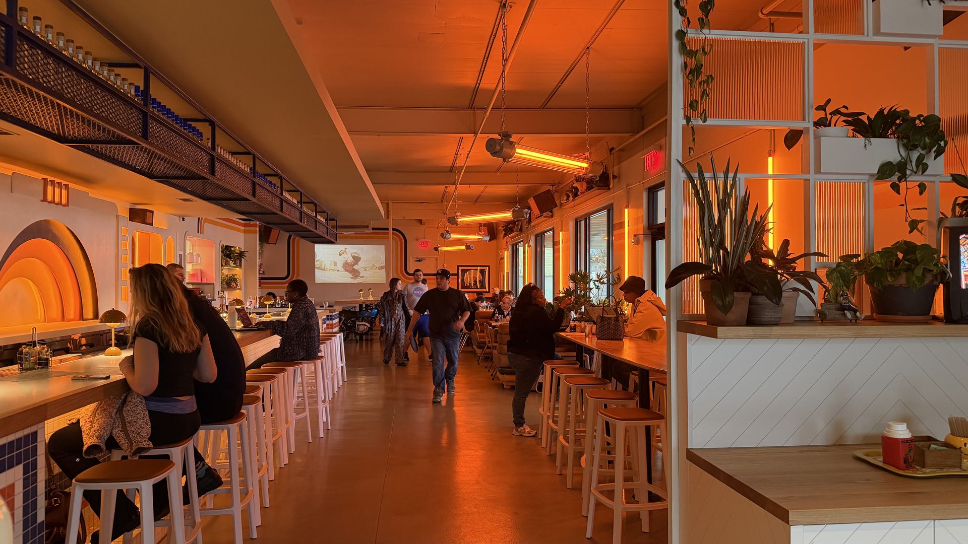 Interior of a warmly lit cafe with orange neon lights, white bar stools, patrons seated at counters and tables, and green plants on shelves and tables.