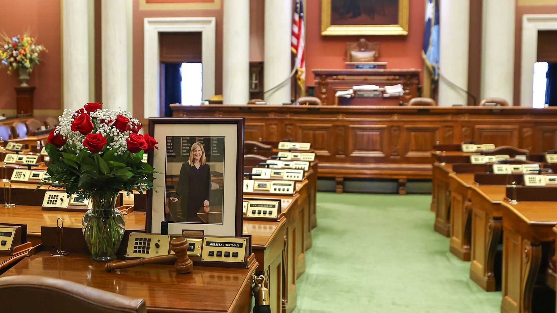 A jar of white and red flowers on a polished wood desk in a legislative chamber with a photograph memorializing former Minnesota House Speaker Melissa Hortman
