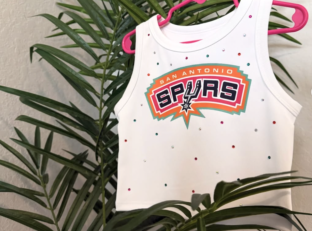 White San Antonio Spurs tank top with a large colorful logo and multicolored rhinestones, hanging on pink plastic hangers in front of green houseplants.