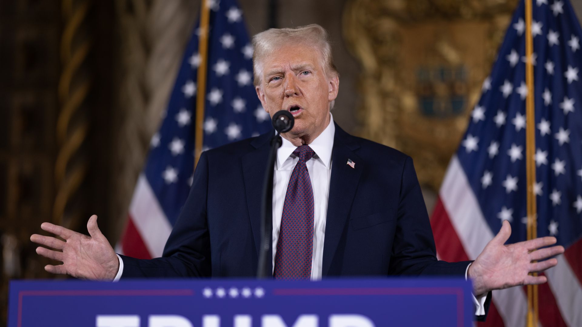 Donald Trump, wearing a blue suit and speaking into a microphone in front of American flags.