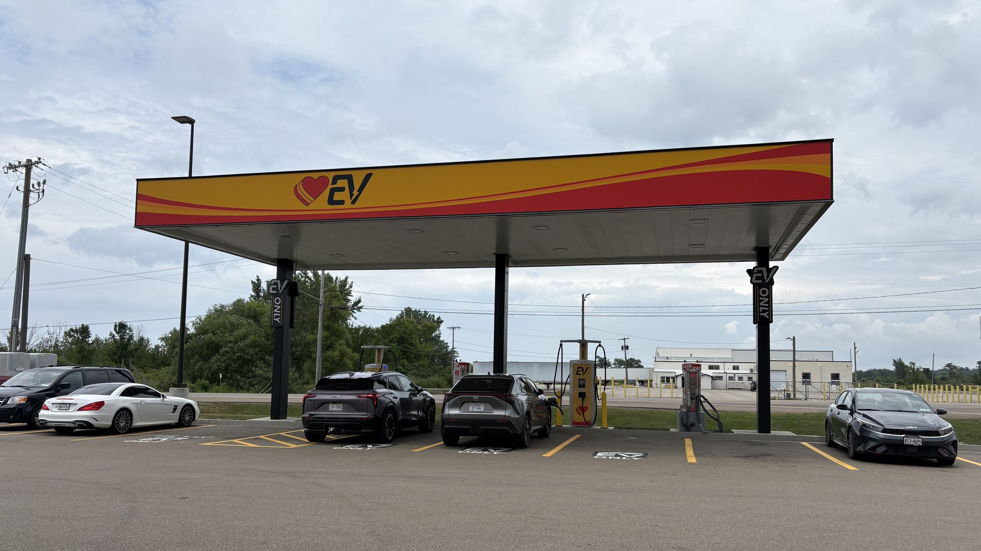 Love's Travel Stop EV charging station with a canopy in yellow and red with an EV logo and heart; several cars parked underneath with a cloudy blue sky.