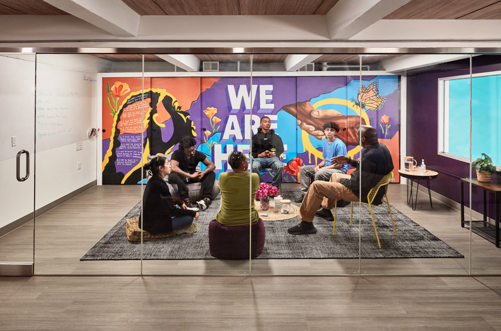 Six diverse people seated in a circle in a room with a colorful mural showing "WE ARE HERE," flowers, and two hands reaching out, visible through glass walls.