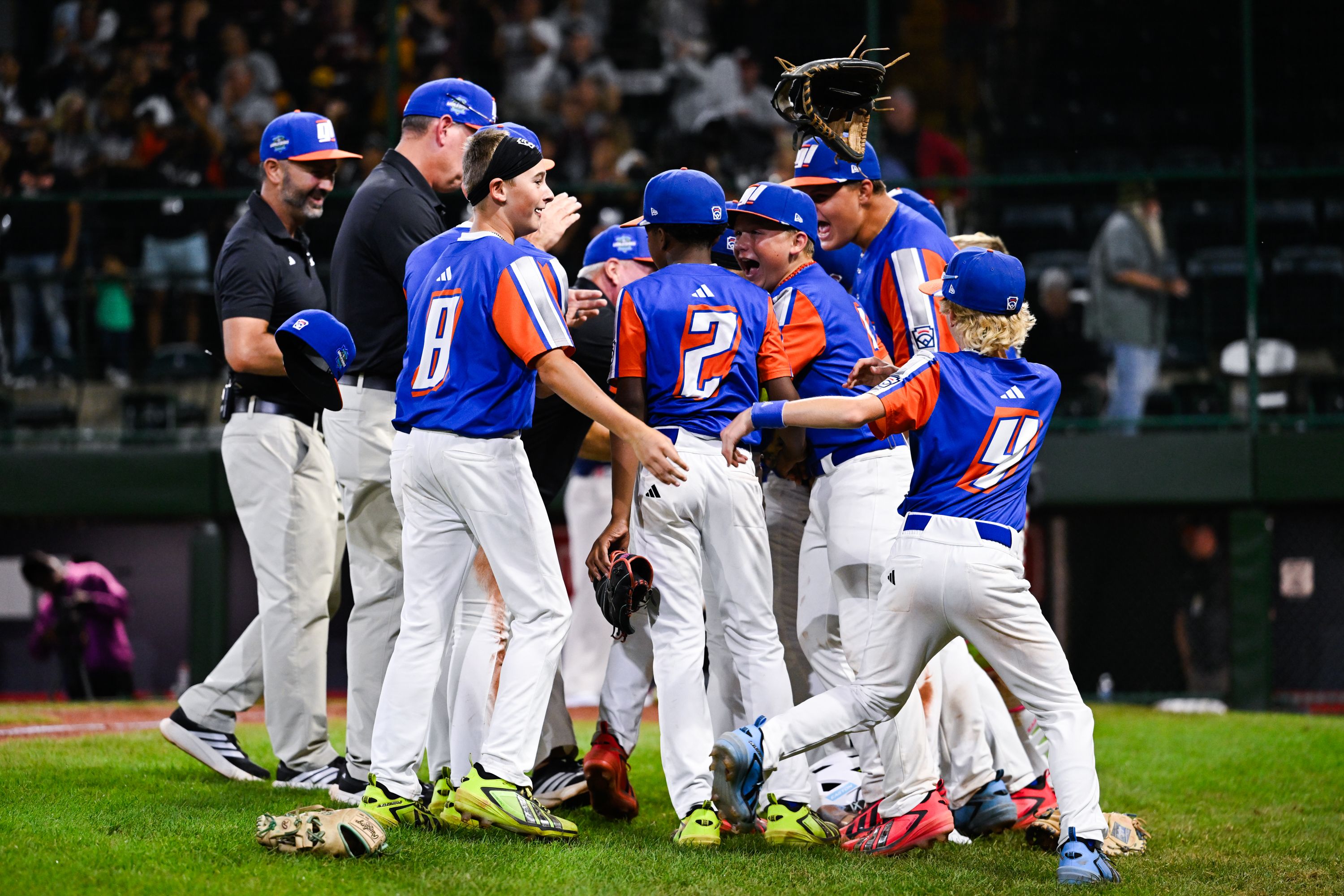 Blue and orange baseball players in a celebratory huddle on the grassy field, white pants and blue caps, raising gloves as a crowd watches from blurred stands.