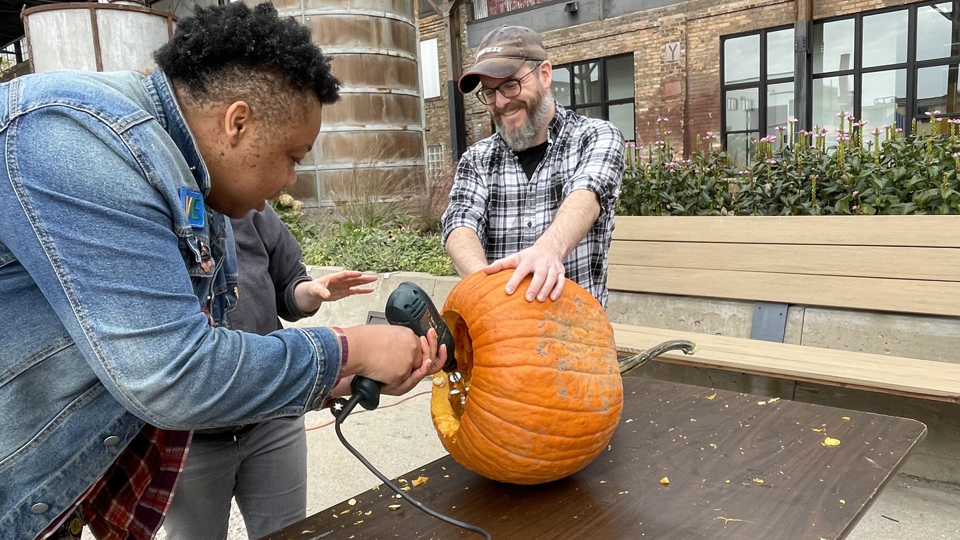 Two men carve a pumpkin at a wooden table outdoors, one in a denim jacket and the other in a plaid shirt and cap. A power tool and cord sit on the table; industrial brick backdrop with a rusted tank.